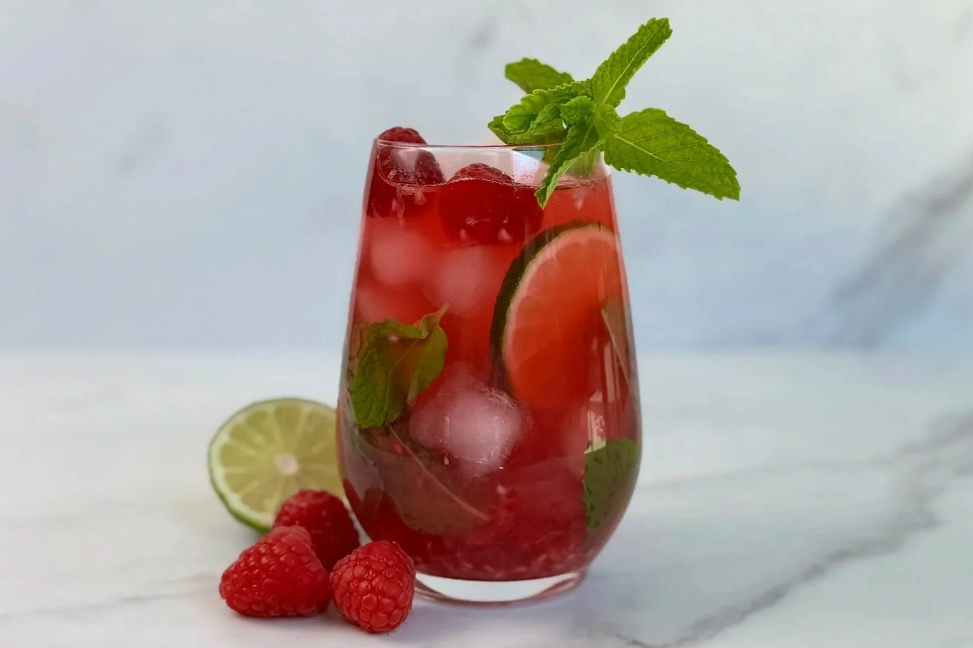 A glass of Raspberry Mojito Mocktail that is garnished with lime wheels and fresh mint, with three raspberries and a lime wedge resting on the countertop.