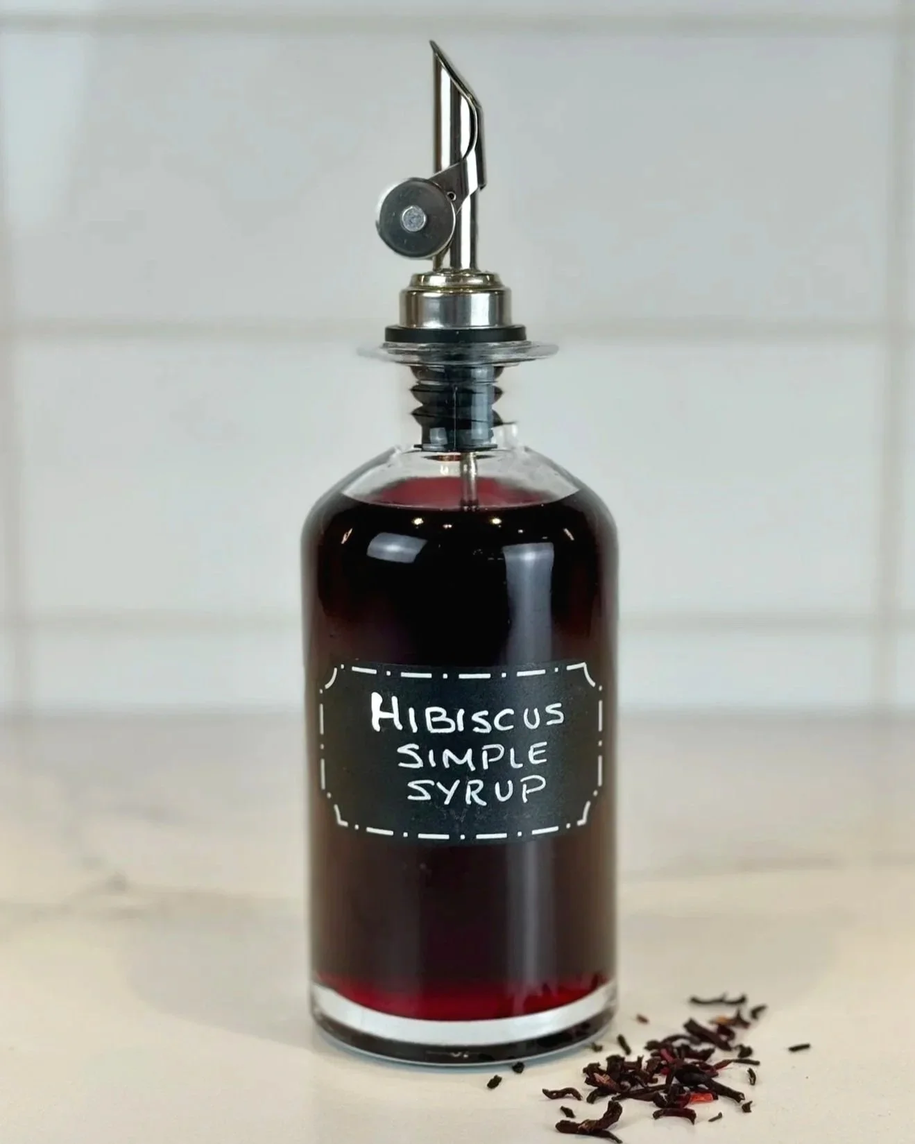 A bottle of Hibiscus Simple Syrup sitting on a marble countertop with a few dried hibiscus flowers on the counter.