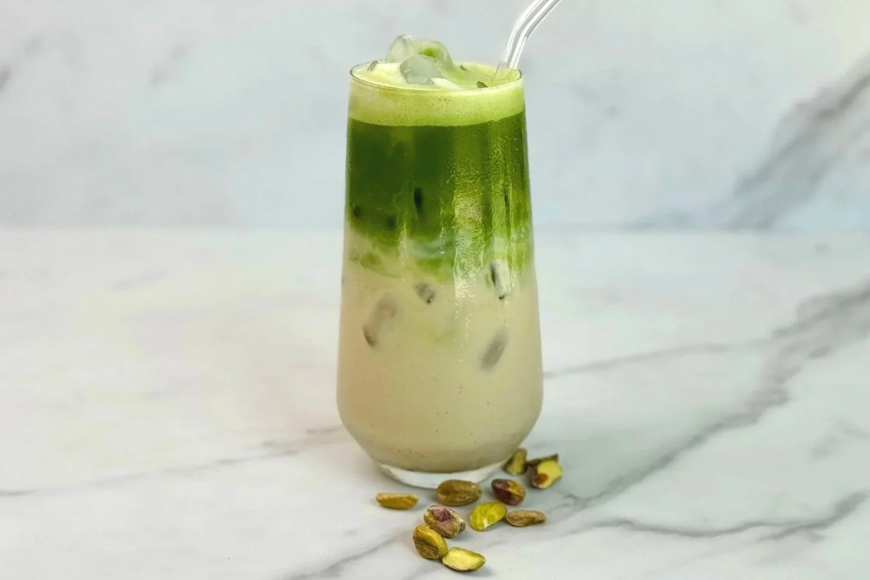 A tall glass of iced Double Pistachio Matcha Latte with a glass straw, and a few pistachio nuts scattered on the counter.