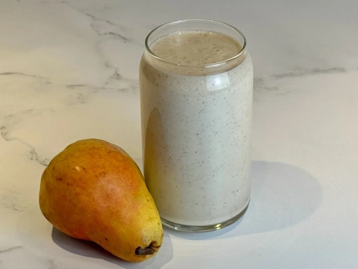 A glass of High Protein Pear Ginger Smoothie sitting on a counter next to a fresh Bartlett pear.