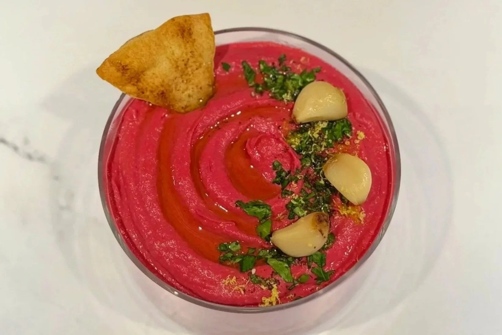 A glass serving bowl of Red Beet Roasted Garlic Hummus, garnished with chopped parsley, Za'Atar, lemon zest, three roasted garlic cloves. A single pita chip is dipped into the hummus.