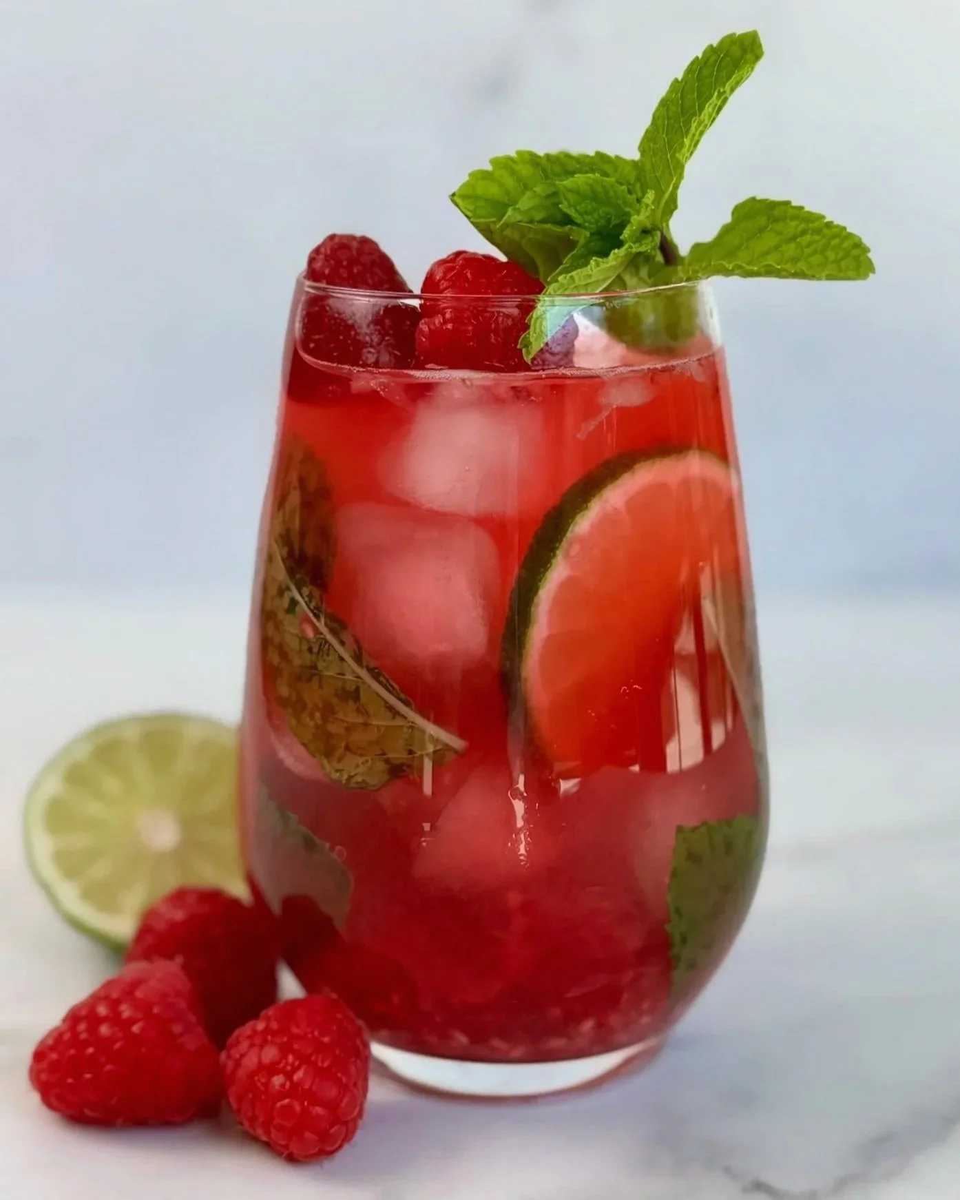 A glass of Raspberry Mojito Mocktail that is garnished with lime wheels and fresh mint, with three raspberries and a lime wedge resting on the countertop.