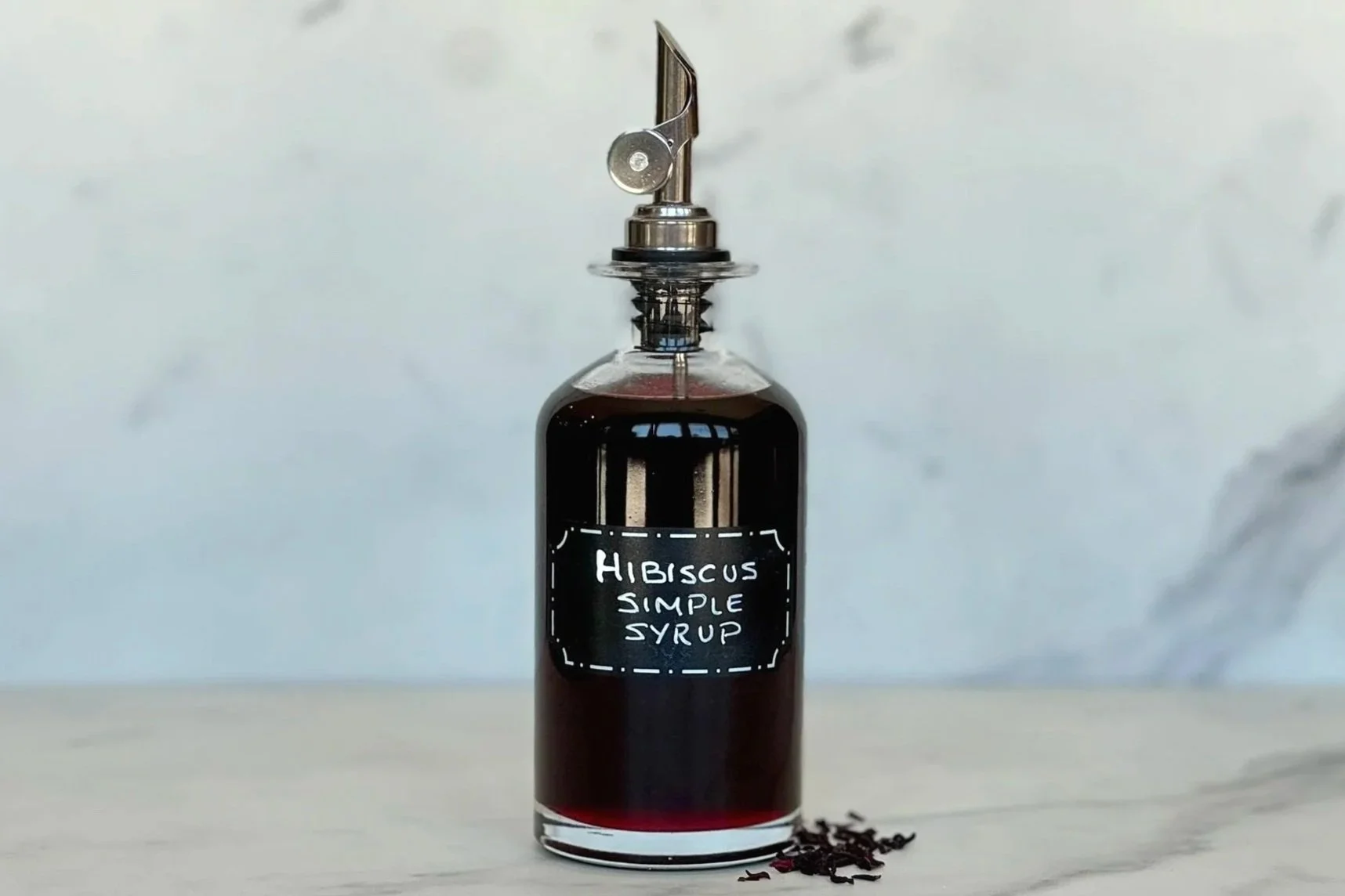 A bottle of Hibiscus Simple Syrup sitting on a marble countertop with a few dried hibiscus flowers on the counter.
