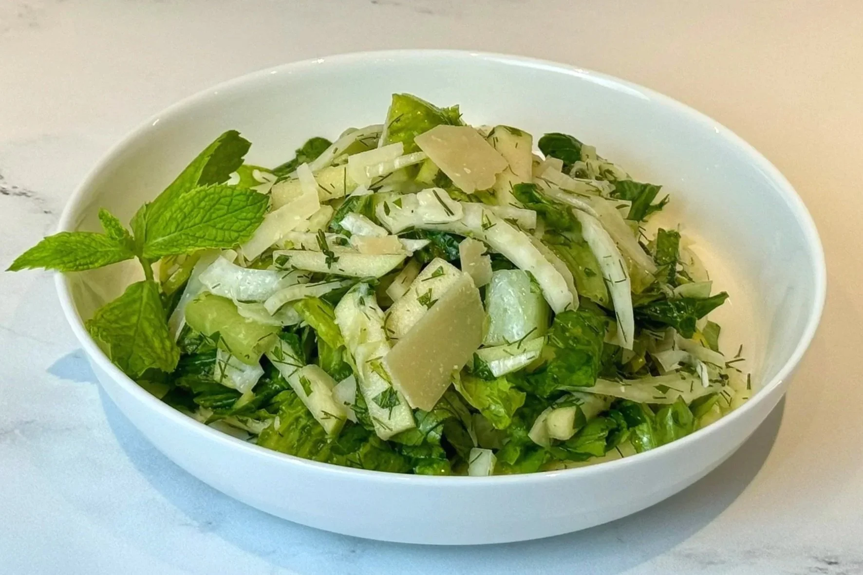 A plate of Herby Fennel & Green Apple Salad garnished with fresh sprig of mint and shaved Parmigiano Reggiano cheese.