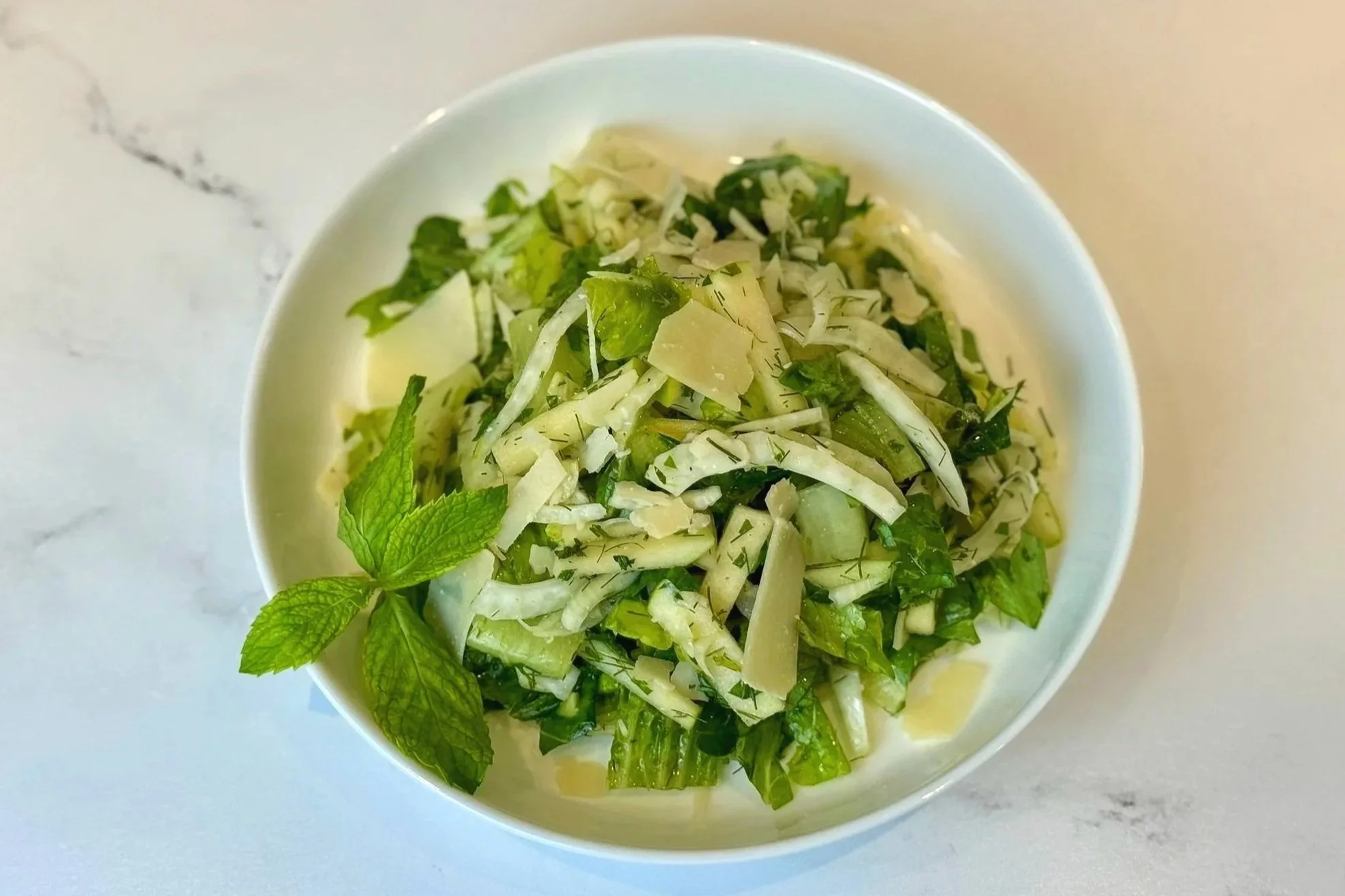 A plate of Herby Fennel & Green Apple Salad garnished with fresh sprig of mint and shaved Parmigiano Reggiano cheese.