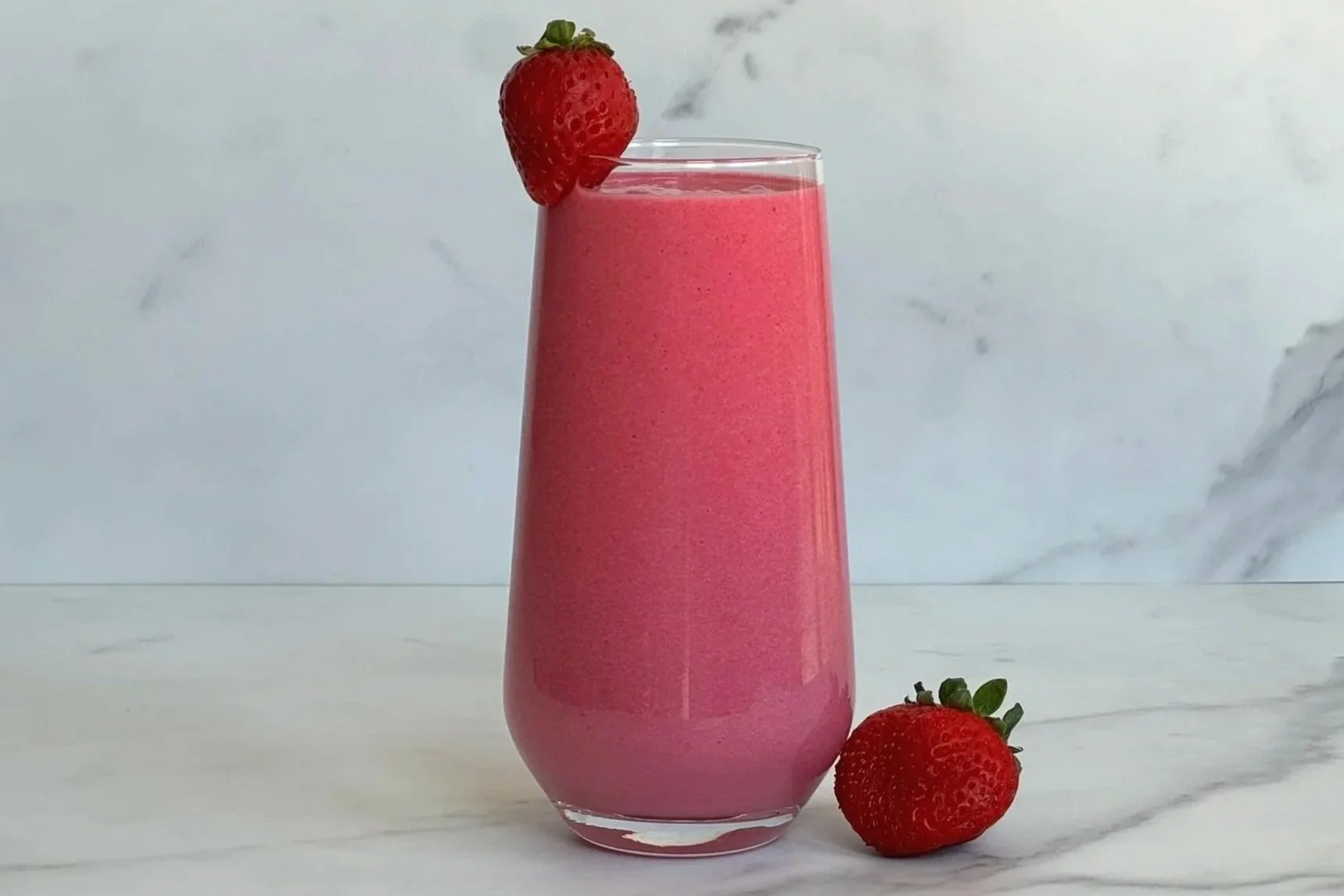 A tall glass of  Strawberry Banana Beet Protein Smoothie, garnished with a strawberry on the glass rim, and one strawberry at the base of the glass.