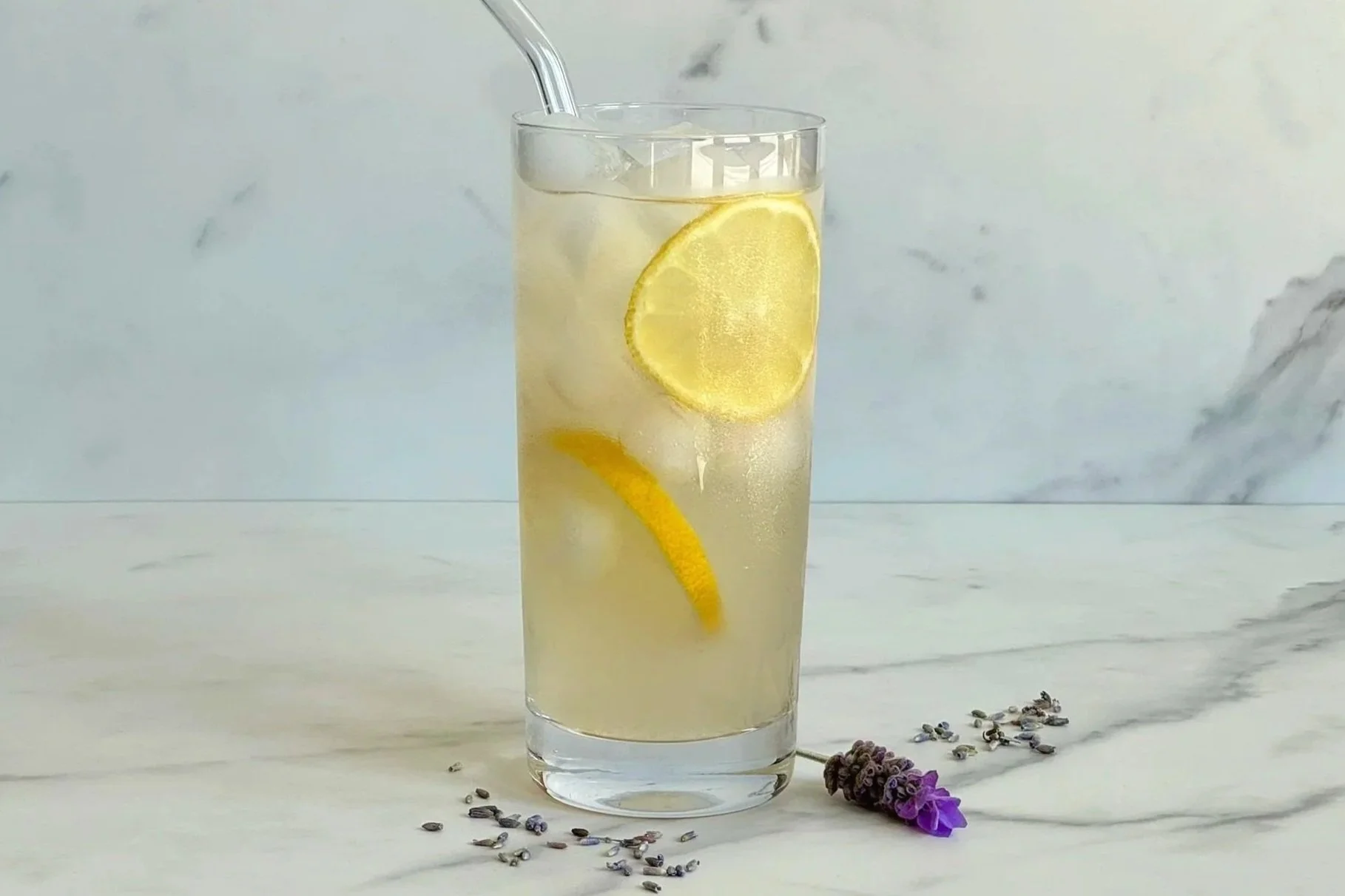 A tall glass of Lavender Lemonade with lemon slices floating in the glass, and a sprig of lavender on the counter along with a few dried lavender buds.