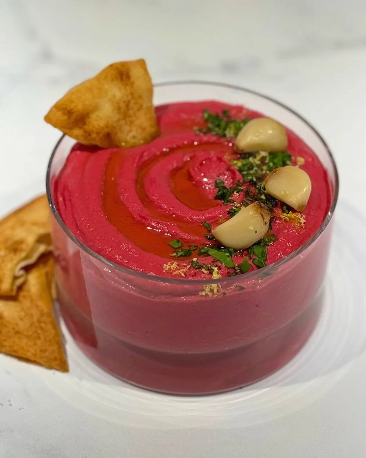 A glass serving bowl of Red Beet Roasted Garlic Hummus, garnished with chopped parsley, Za'Atar, lemon zest, three roasted garlic cloves. A single pita chip is dipped into the hummus, and two pita chips on the counter.
