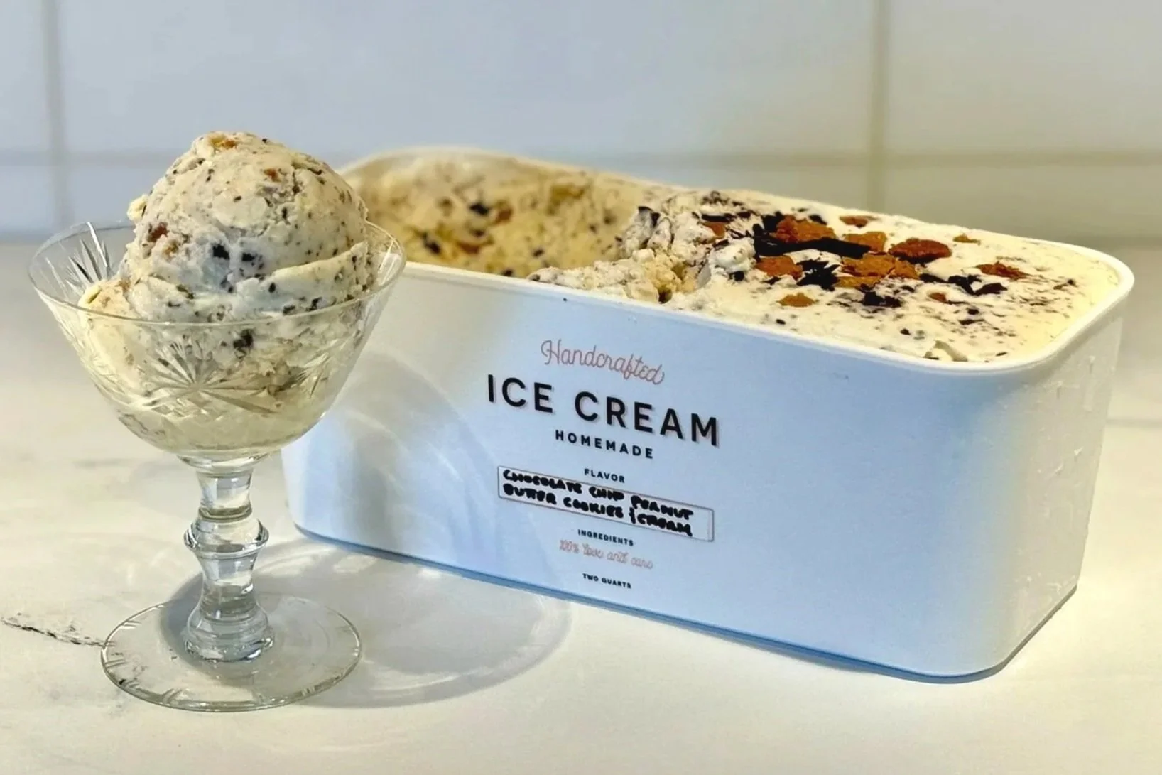 A large scoop of Chocolate Chip Peanut Butter Cookies & Cream Ice Cream served in a crystal goblet, alongside a rectangular container filled with this ice cream.
