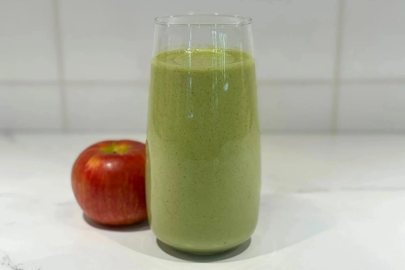 A tall glass of Apple Kale Smoothie sitting on a marble counter with a whole red apple resting beside the glass.