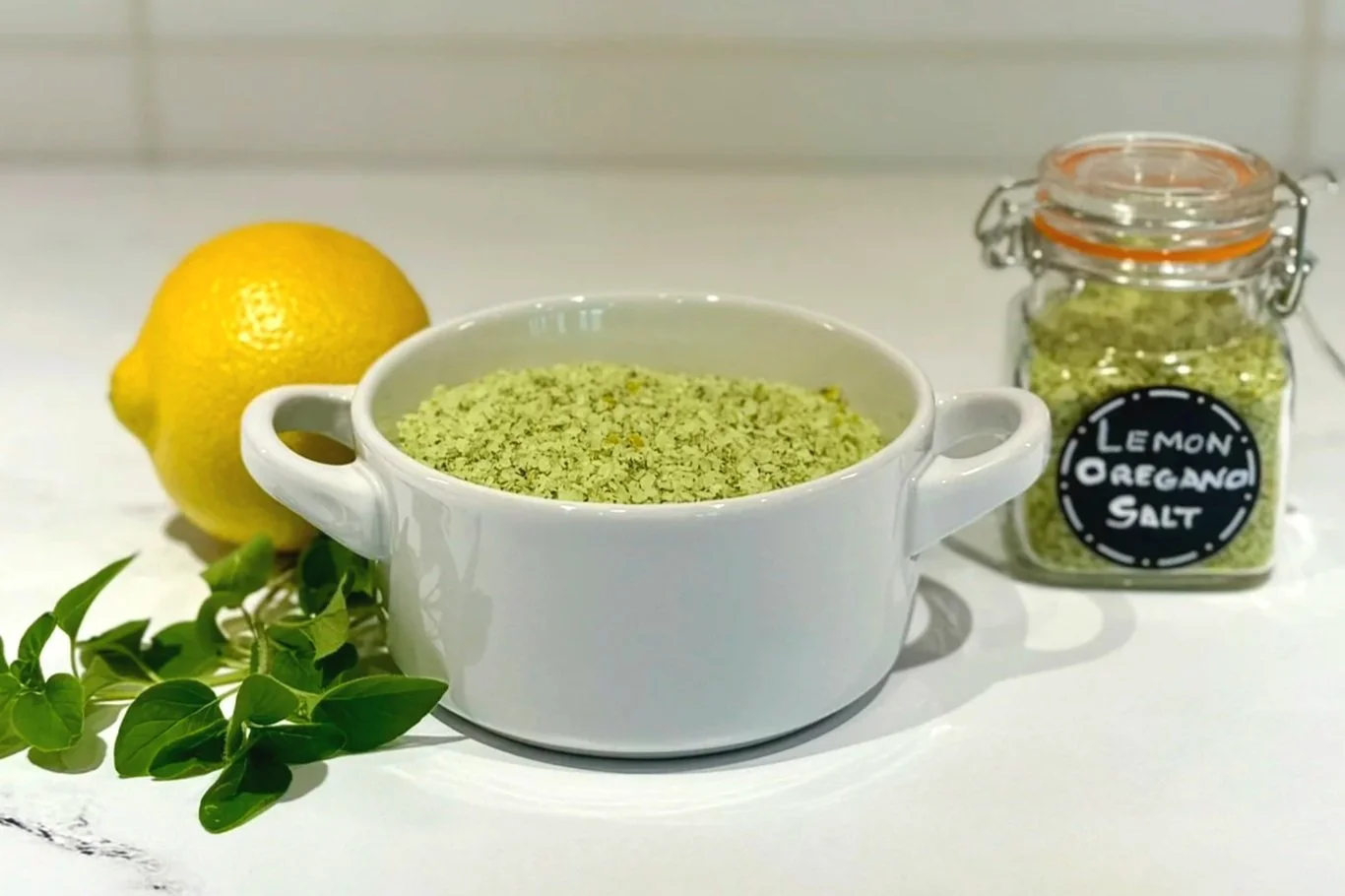 A white Le Creuset salt keep and a small Spice Jar with a flip lid filled with Lemon Oregano Salt . A whole lemon and a sprig of fresh oregano are resting on the counter next to them.