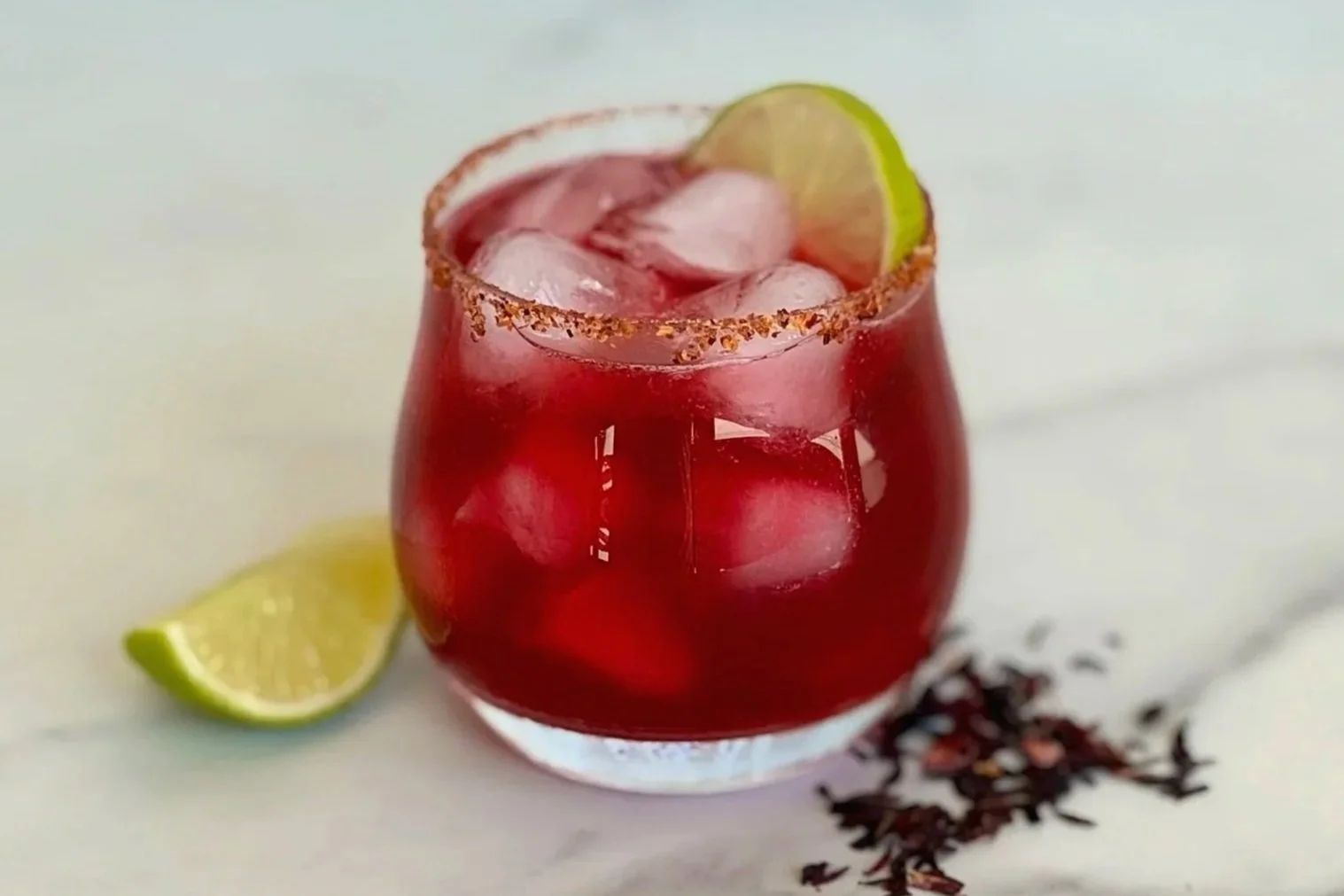 A cold cocktail glass of Hibiscus Mezcal Margarita that is garnished with a Tajin rim and lime slice. A wedge of lime and some dried hibiscus buds are scattered on the counter.