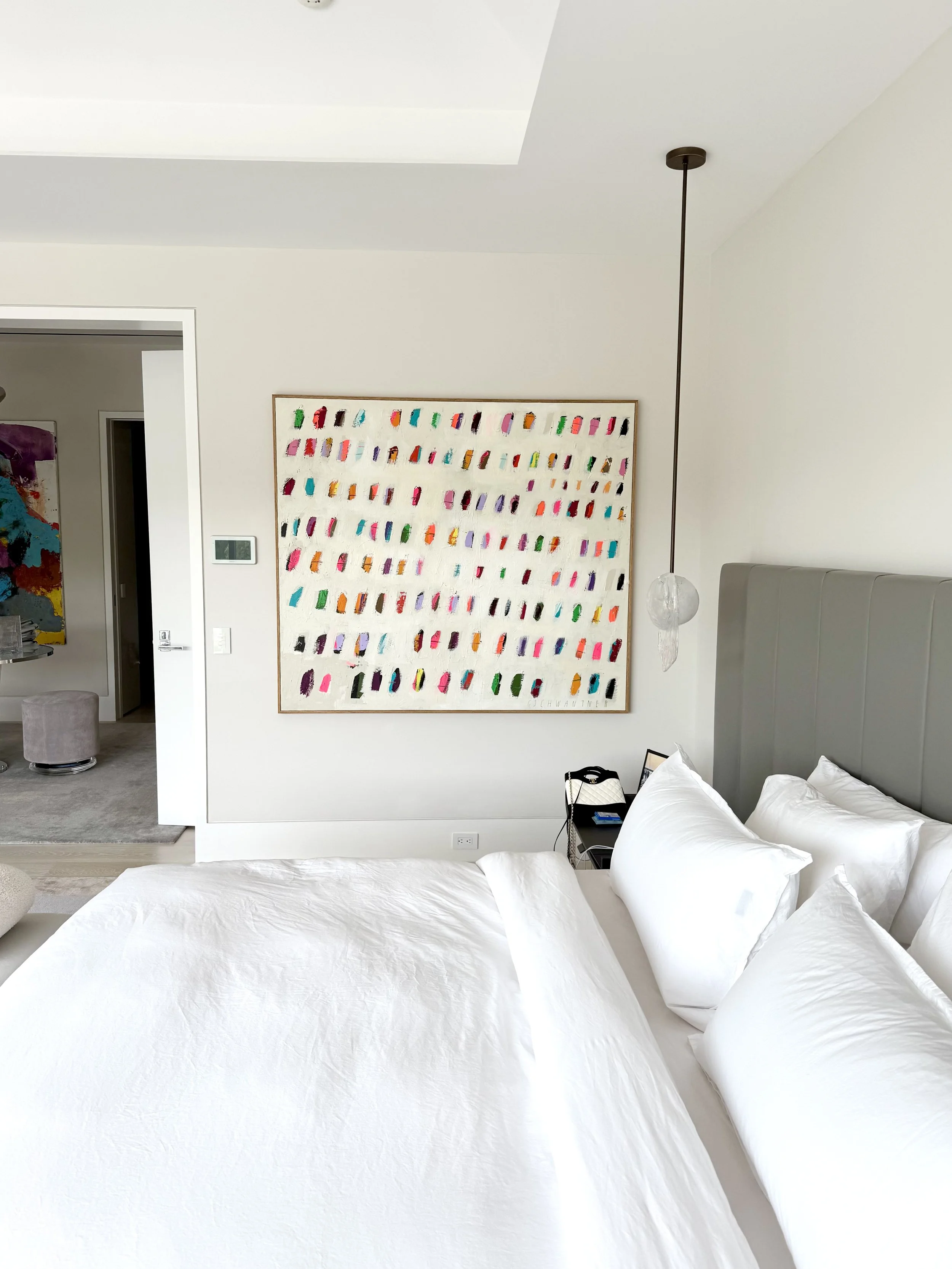 A bedroom with a white bed, pillows, and a nightstand with a phone, against a background of abstract artwork and a hanging light fixture.