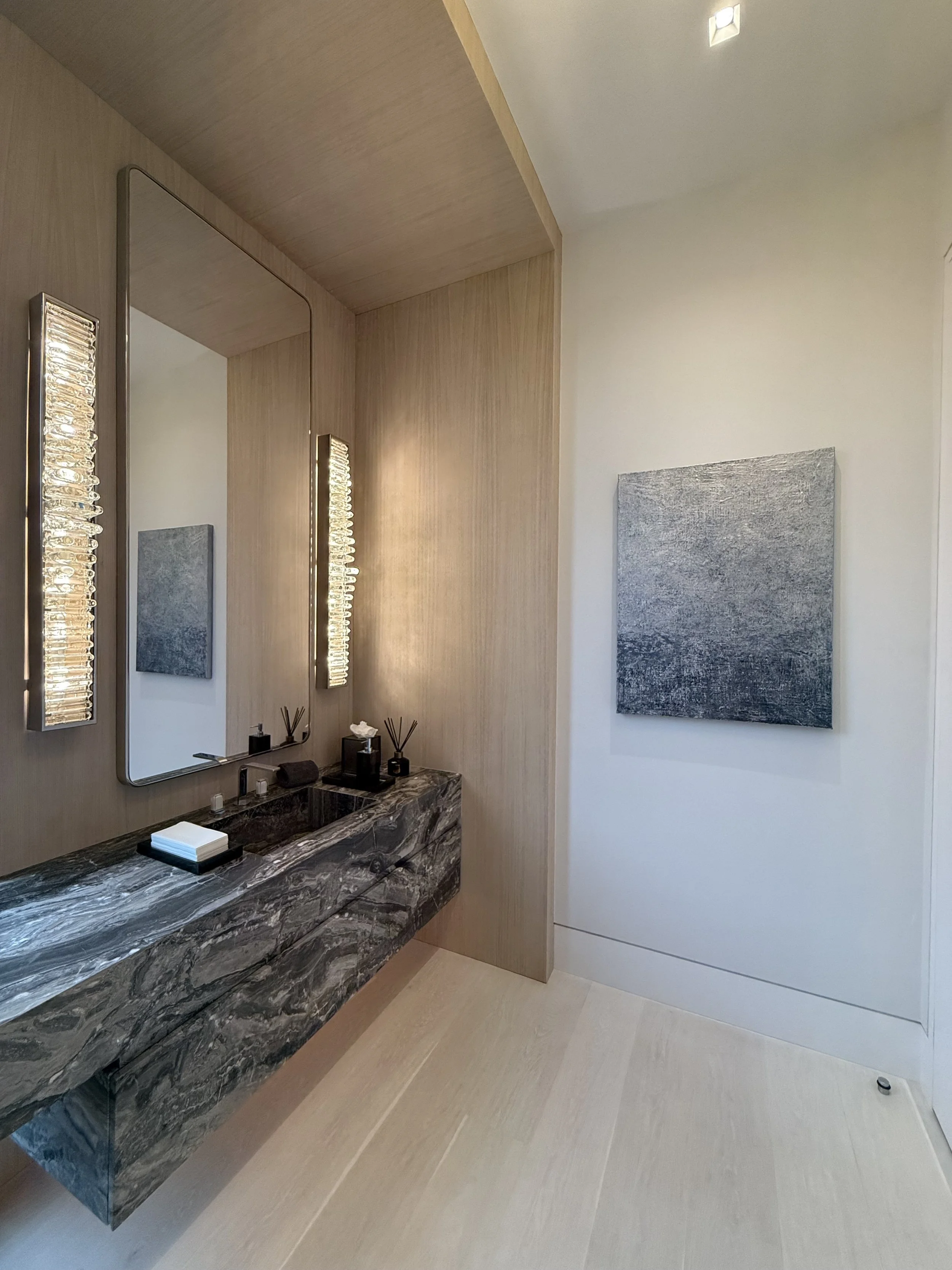 Modern bathroom with black marble countertop, large mirror, textured wall sconces, and a textured gray artwork on a white wall.