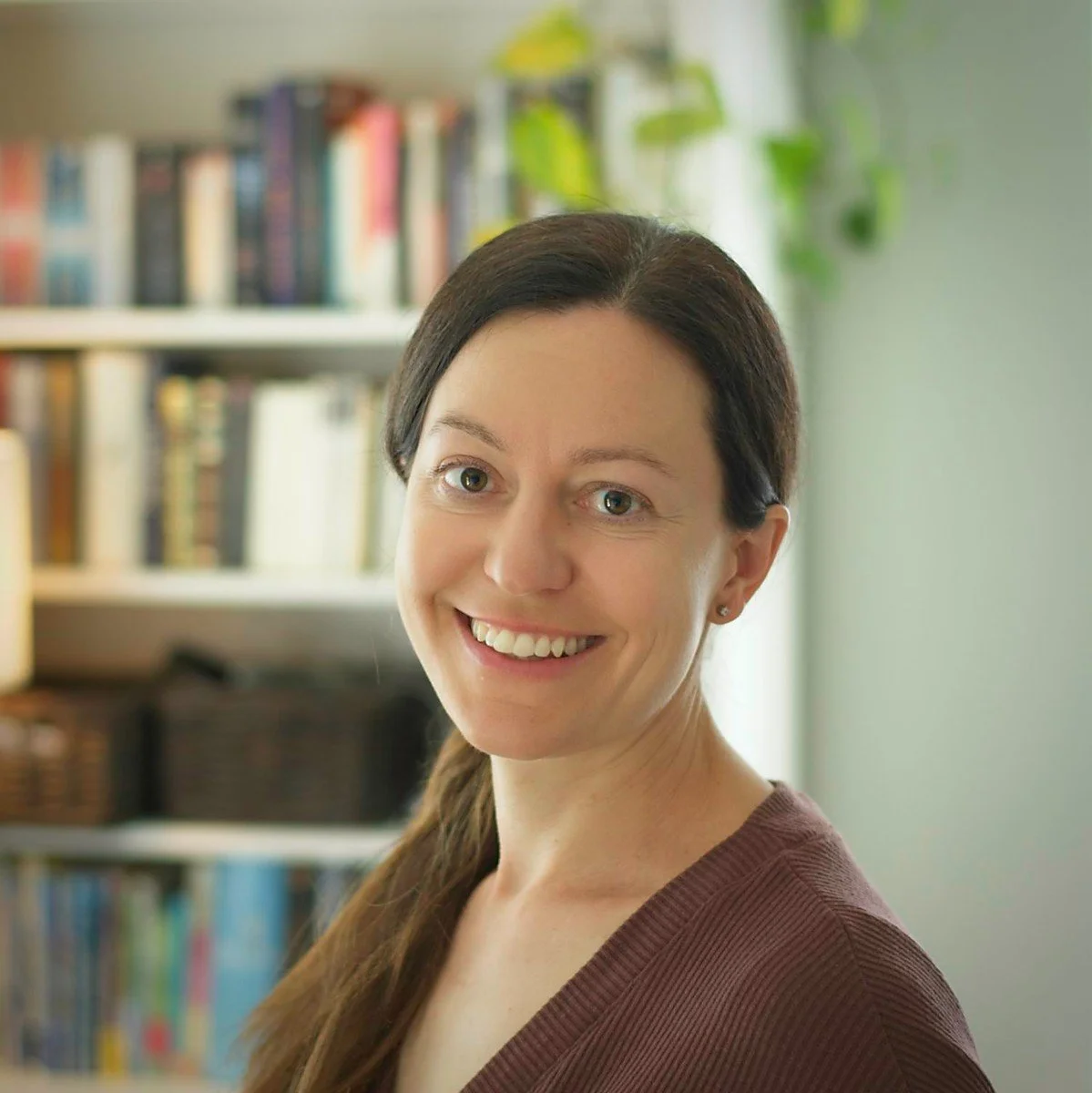 A woman with brown hair and green eyes, smiling, in front of a bookshelf in a bright room.