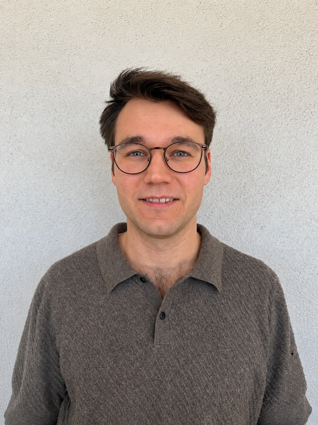 A young man with brown hair, glasses, and light skin smiling in front of a plain white wall.