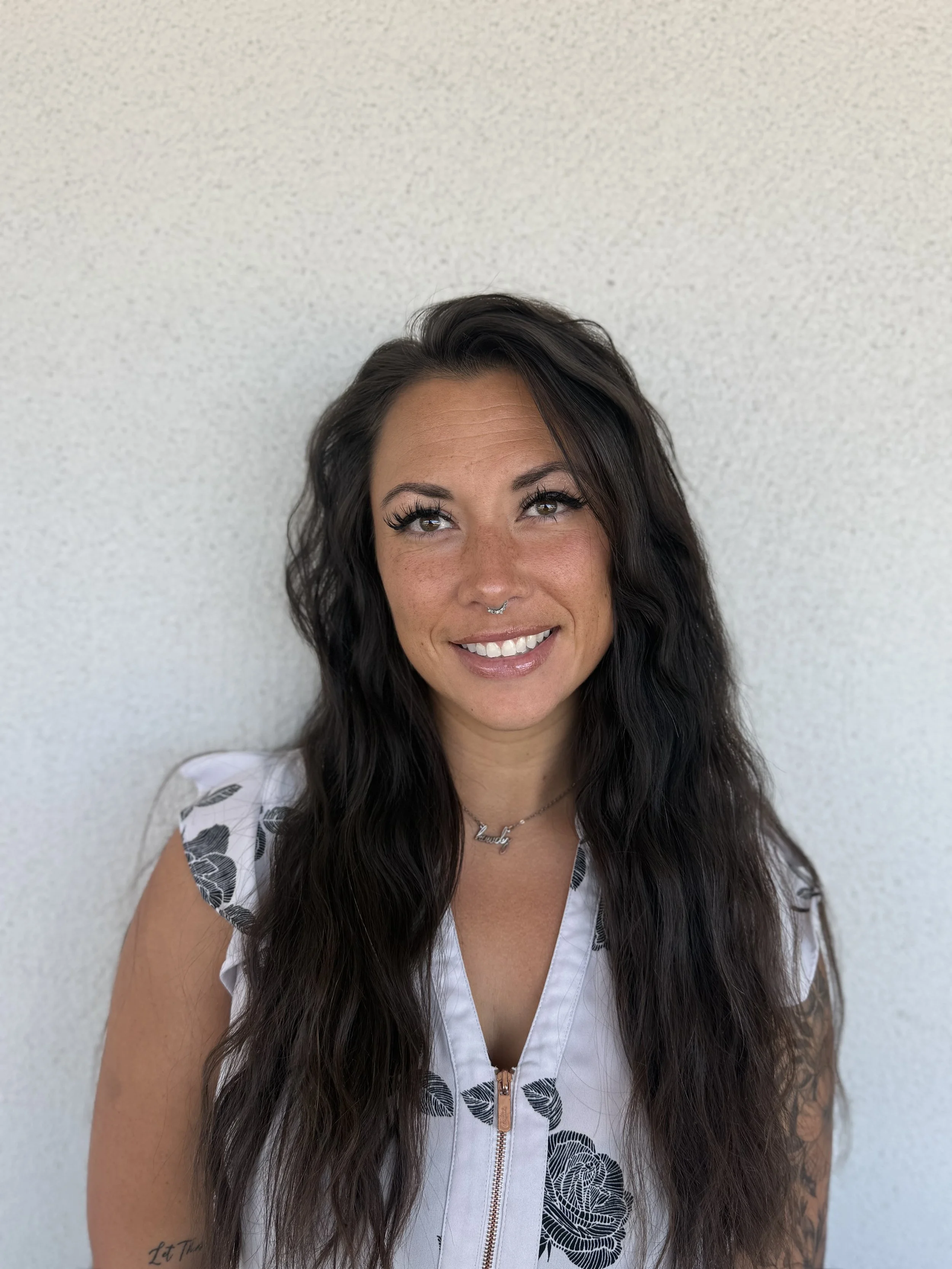 A woman with long dark hair, wearing a white top with black leaf patterns, smiling, standing against a plain textured white wall.