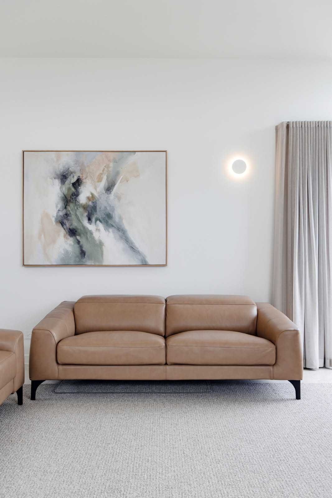 Living room with beige leather sofa, abstract wall art, curtain, and round wall light.