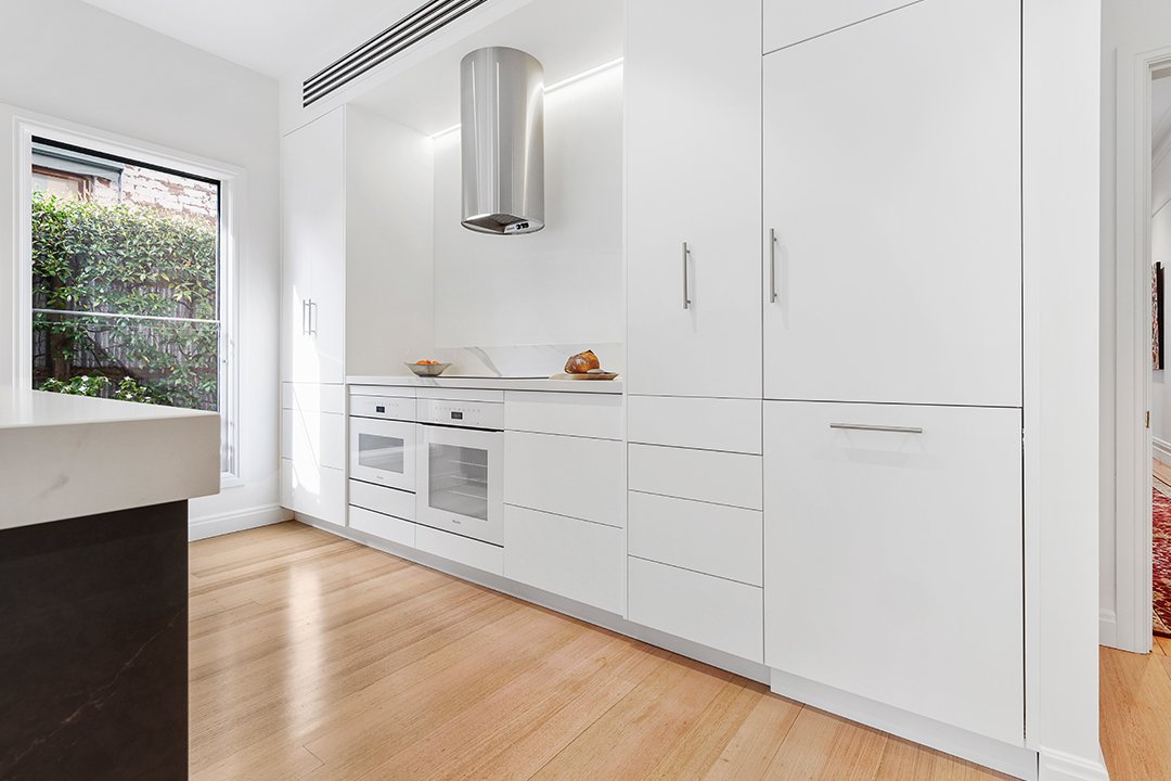 Modern white kitchen with built-in oven, cooktop, stainless steel range hood, cabinets, hardwood floor, and window with greenery outside.