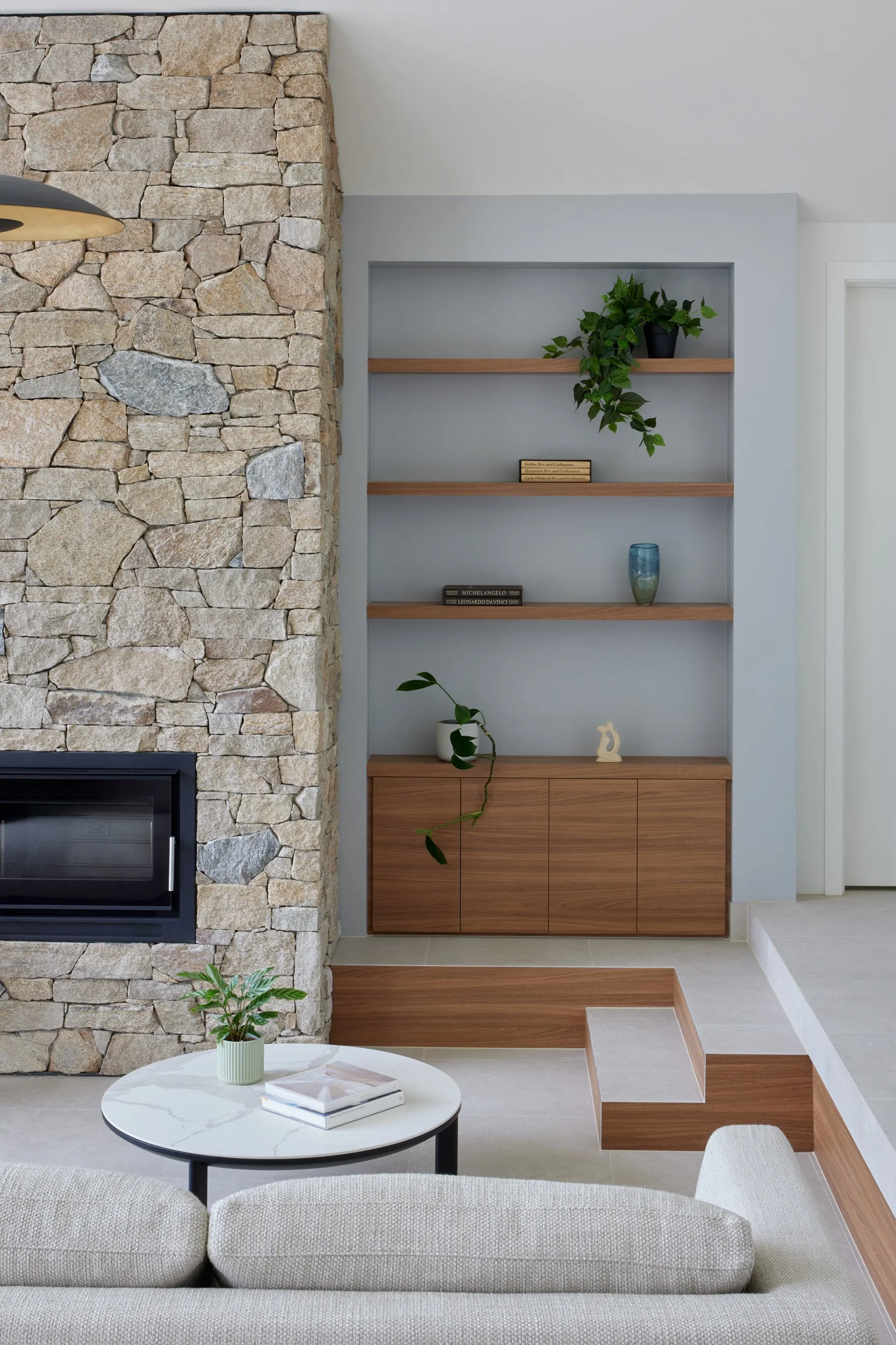 Living room with stone fireplace, built-in wooden shelves with plants and decorative items, and a small round coffee table with books and a potted plant.