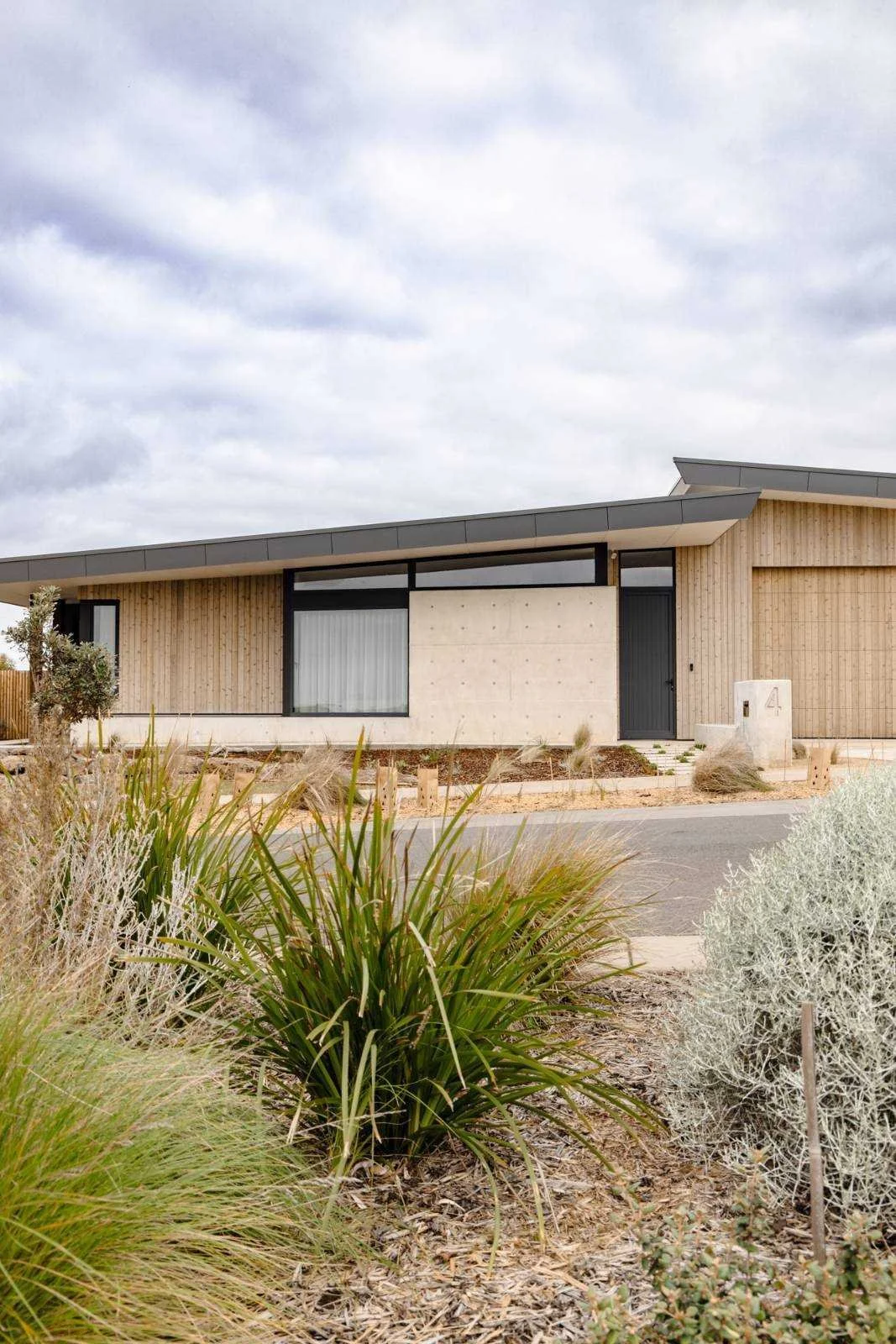 Front view of modern house with wooden and concrete exterior, gray roof, and landscaped garden with various plants and shrubs.