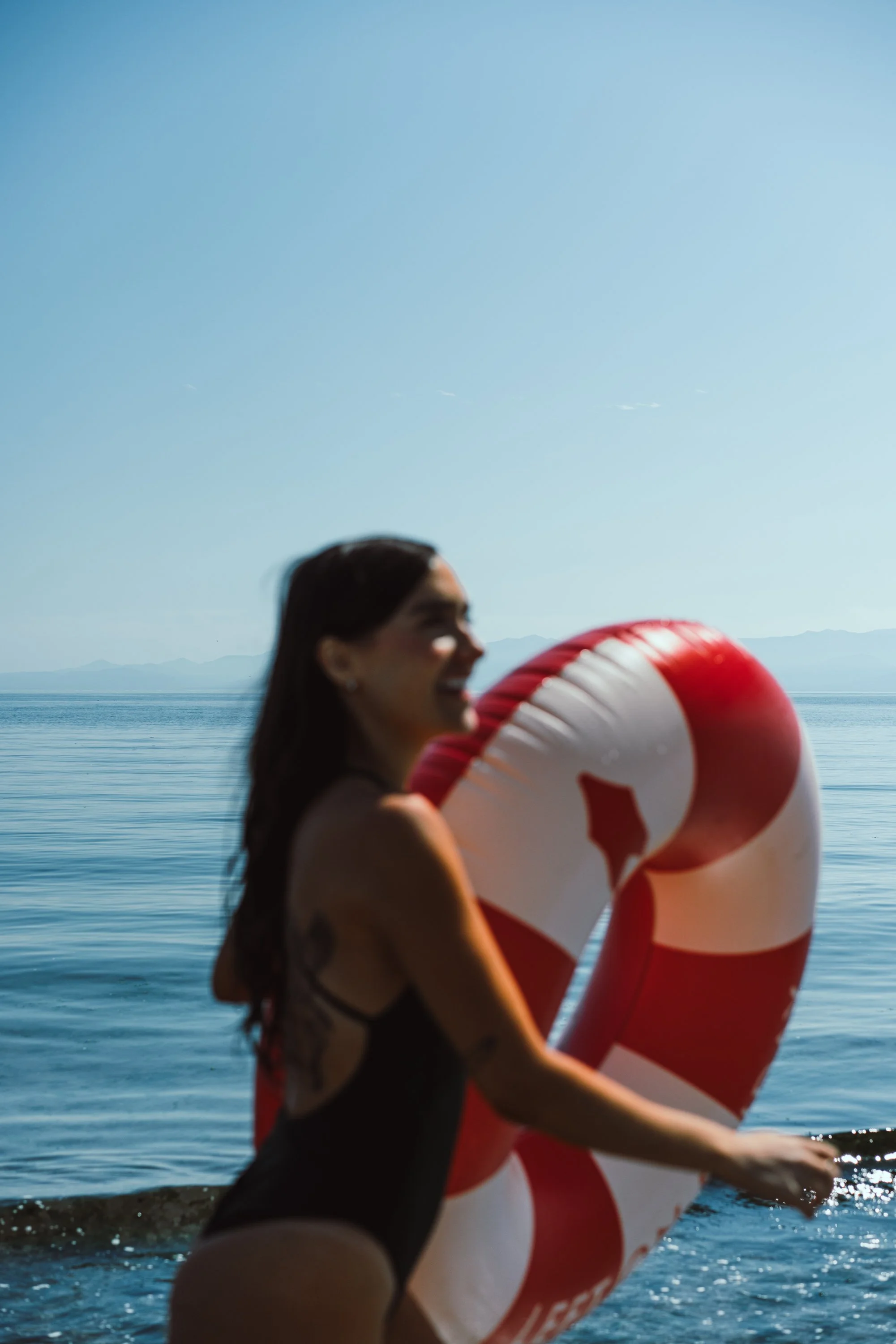 A woman with long dark hair in a black swimsuit holding a red and white life preserver at the beach, with water and distant mountains in the background.