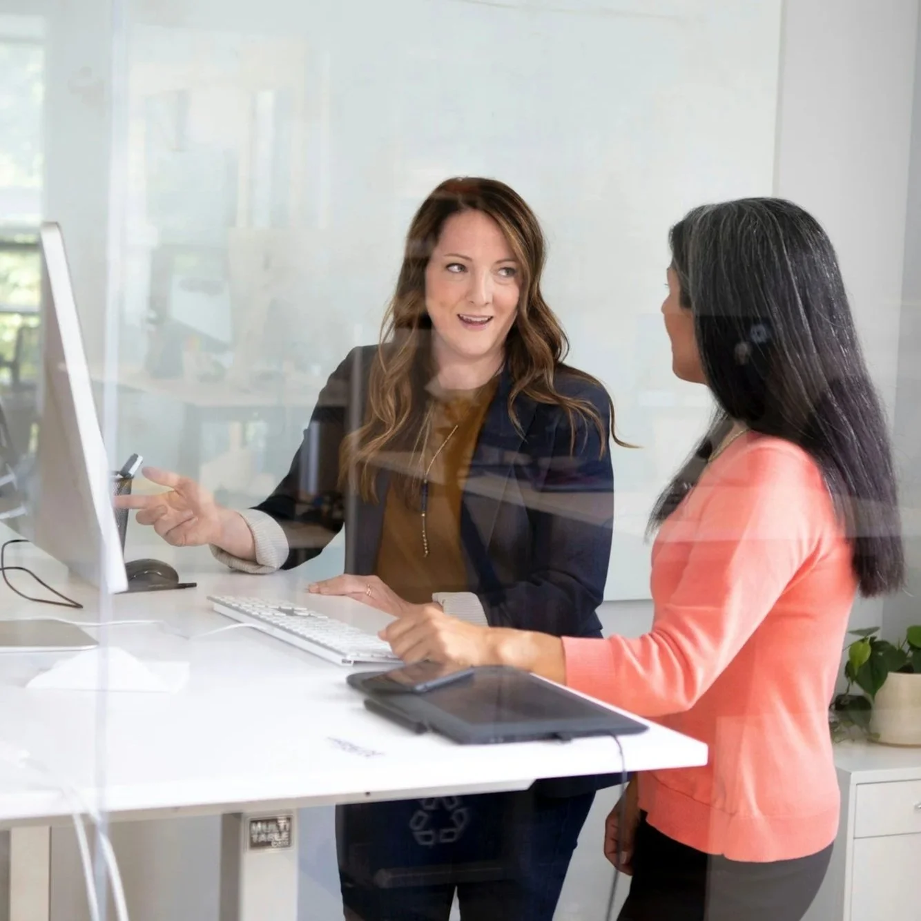 Two women stand at a desk talking in a modern office as one, a Fractional Financial Controller, points at the computer monitor.