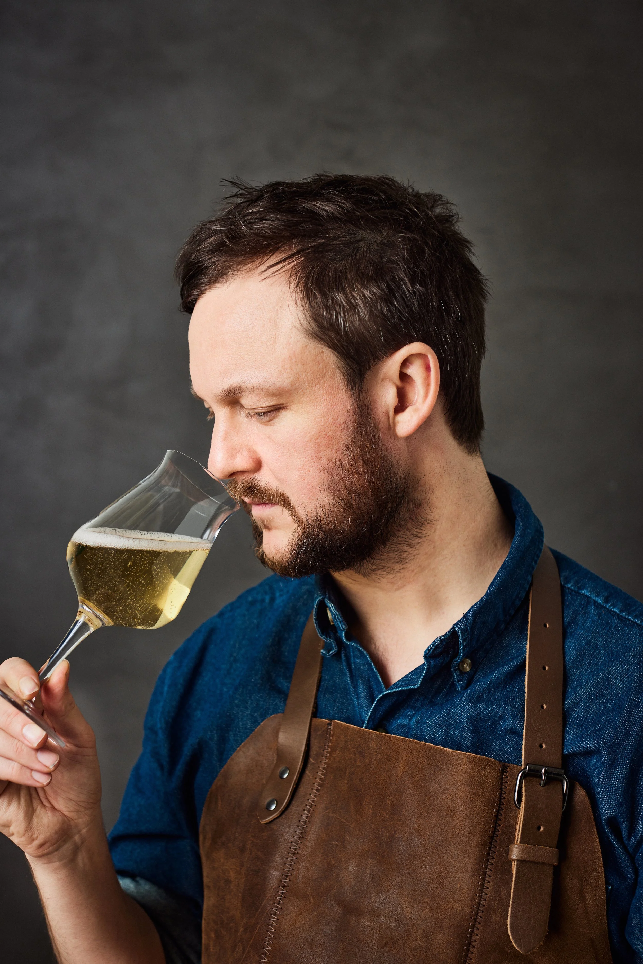 Man smelling a glass of norwegian cider, wearing a denim shirt and a leather apron against a gray background.