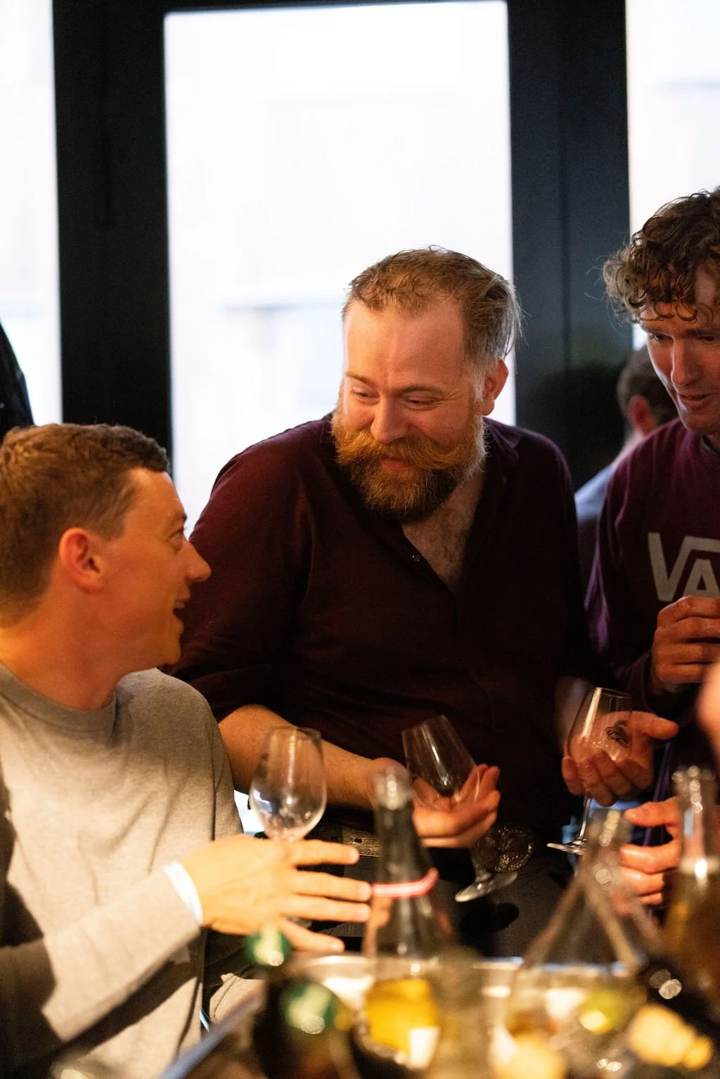 Three men at a social gathering sharing drinks and smiling, with bottles and glasses on the table.
