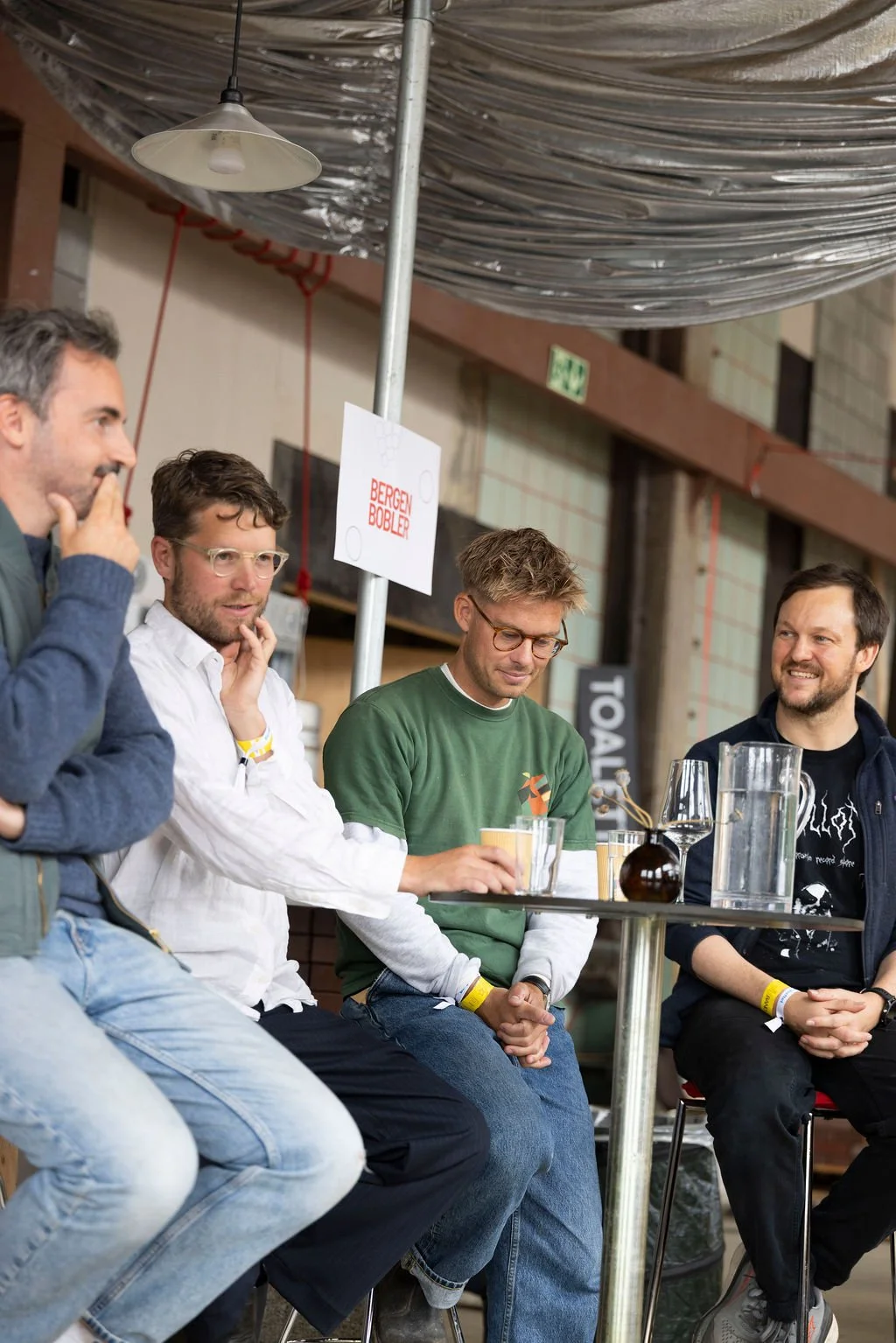 Four men seated at a table in a brewery or bar, engaged in conversation, with drinks and a small vase on the table, and a sign that reads 'BERGEN BOBLER' behind them.