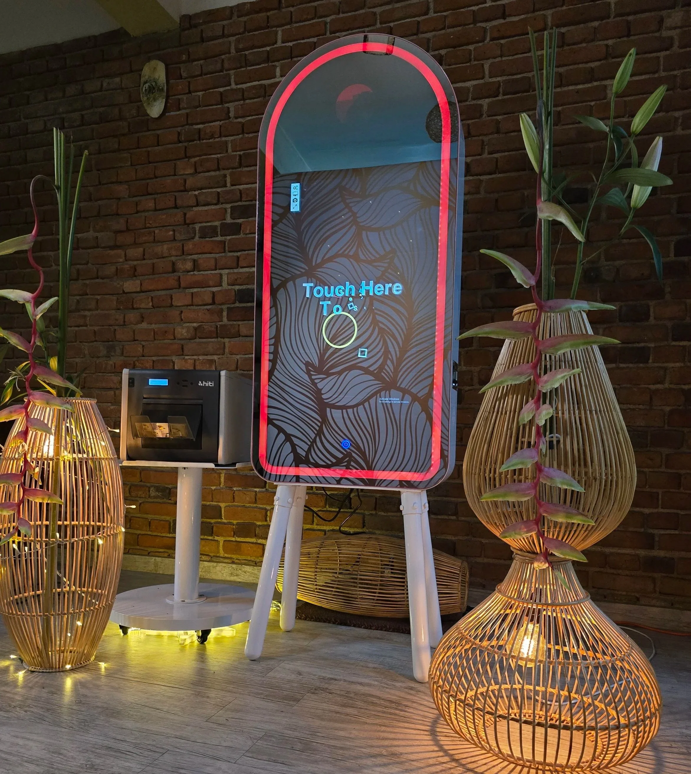 Indoor scene featuring a large digital touchscreen kiosk with a message 'Touch Here To' and a yellow circle, illuminated by warm yellow light. To the right of the kiosk are two wicker lamps and a wicker chair, with a potted plant nearby. The background consists of a red brick wall.