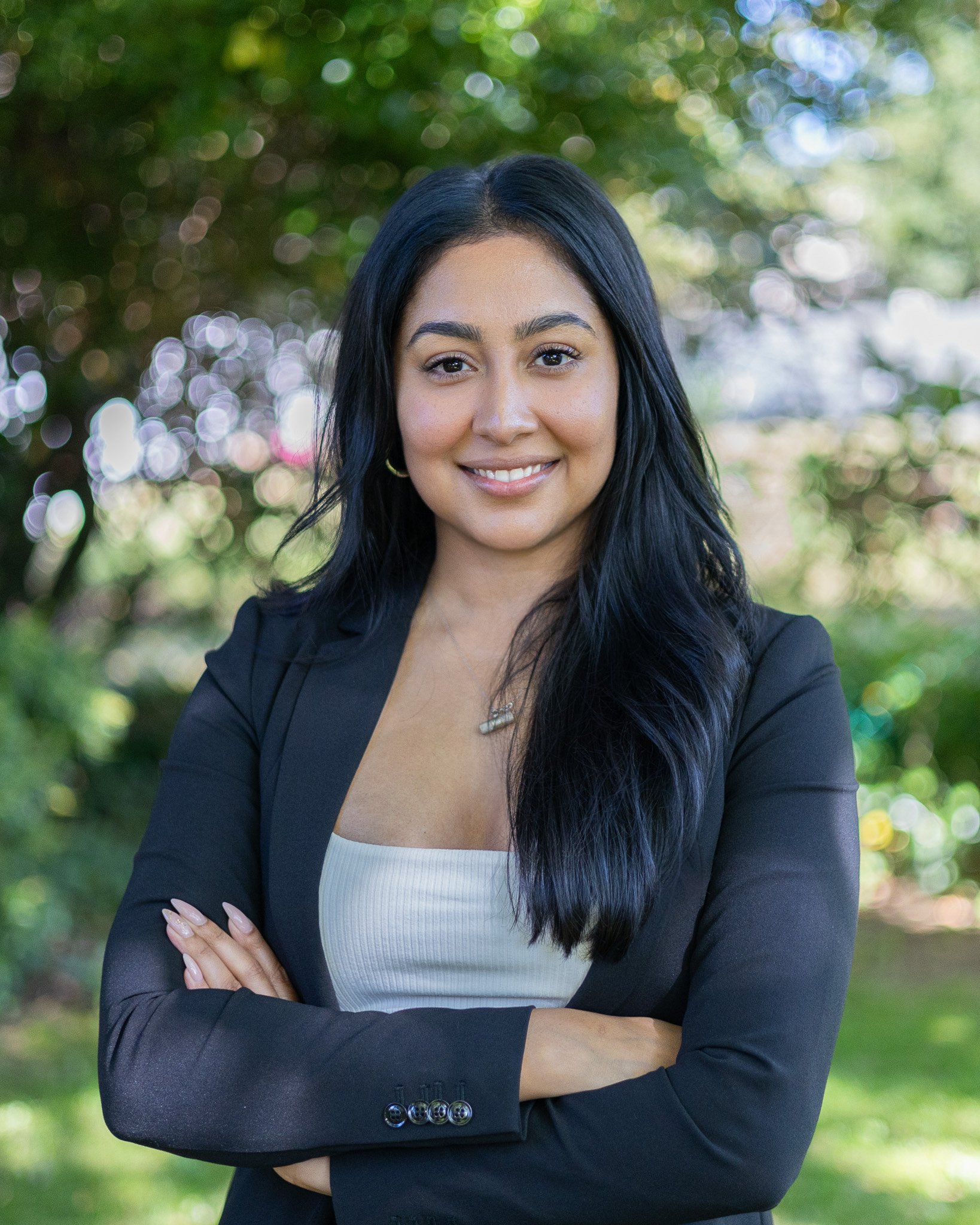 Woman with long black hair, wearing a black blazer and white top, standing outdoors with arms crossed, smiling, green blurred background.
