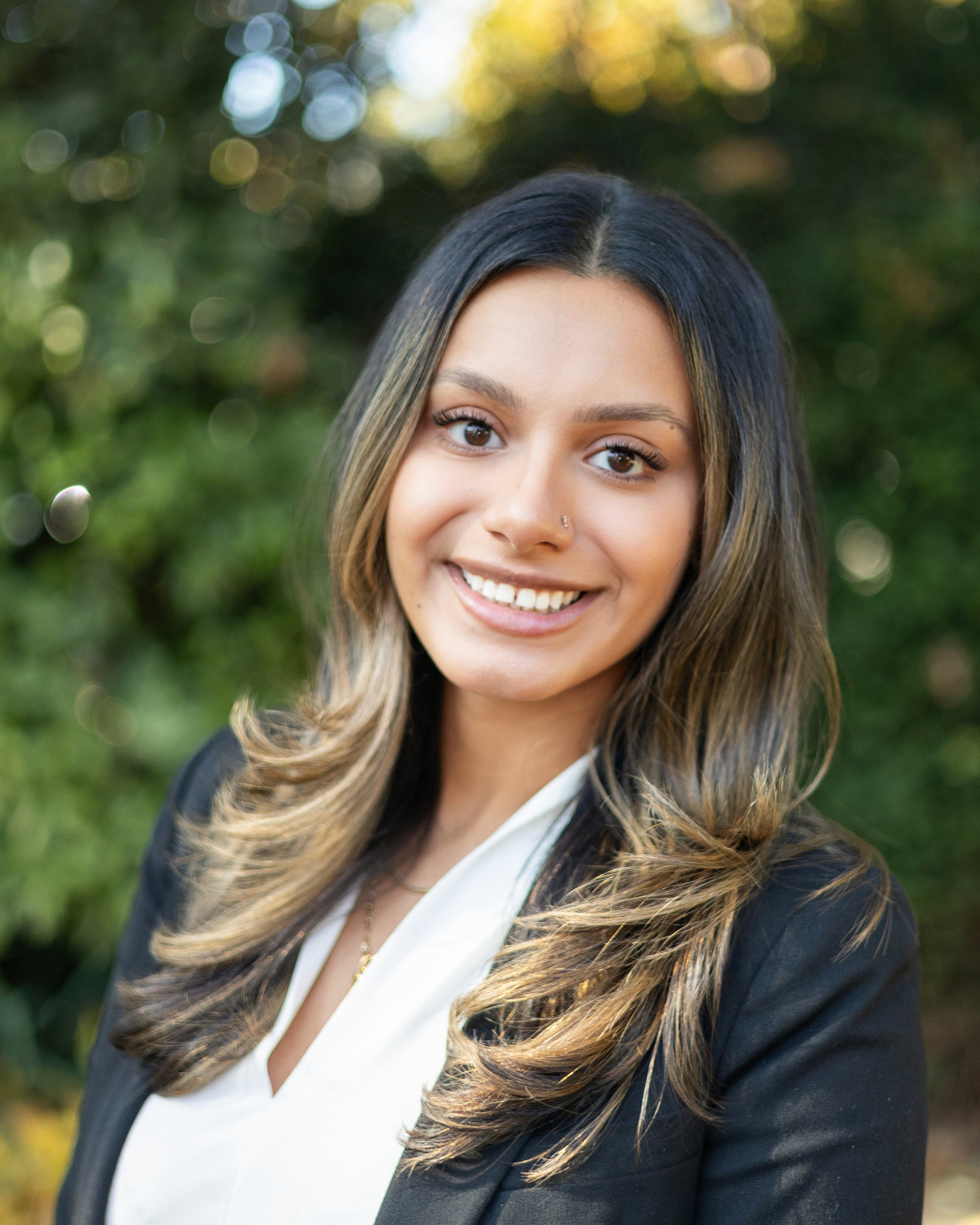 Smiling person with long hair outdoors, wearing a black blazer and white shirt.