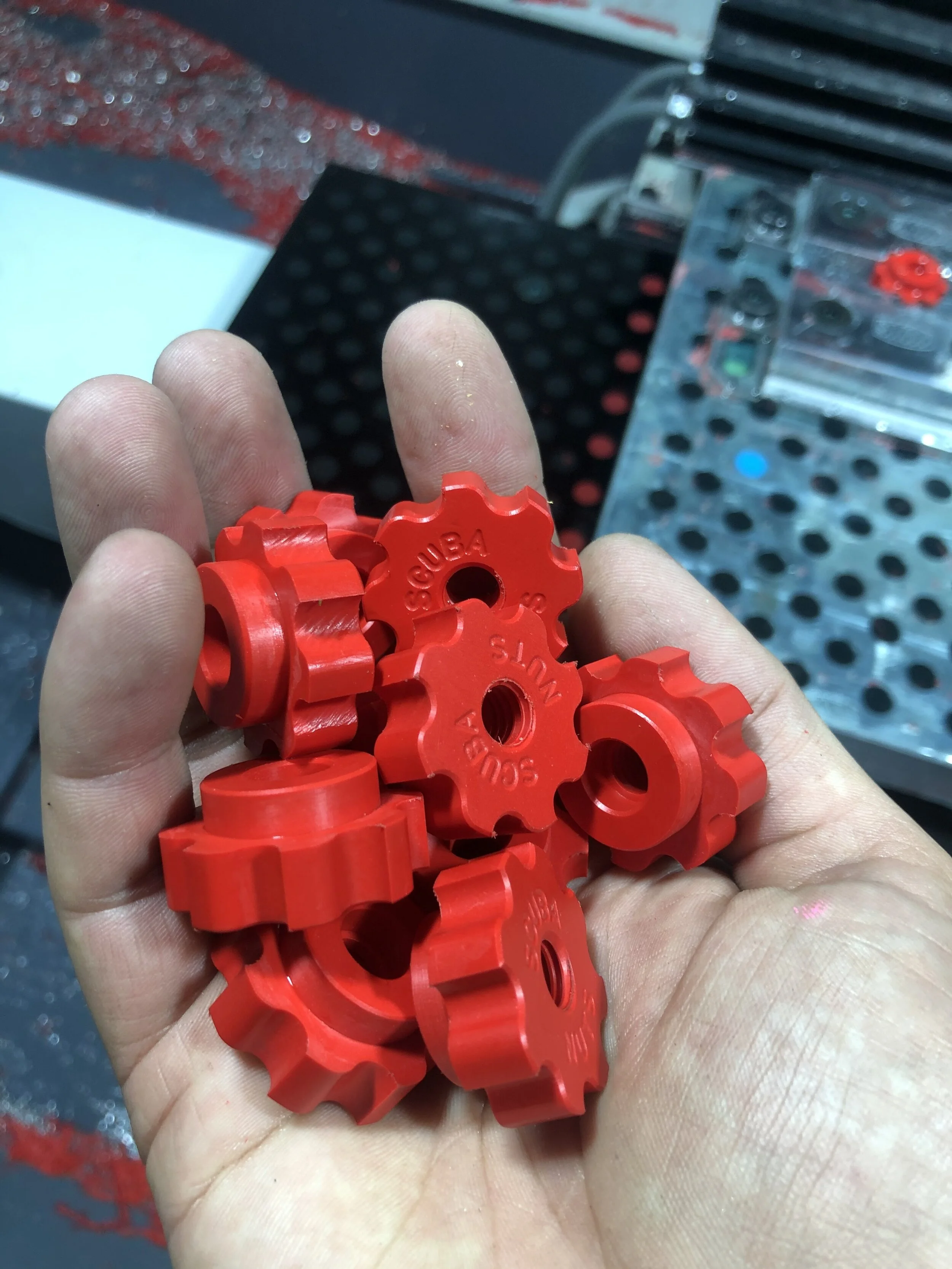 A person's hand holding several red plastic gears with the words 'SOUTHEAST USA' and 'S' engraved on them, with a workshop or manufacturing setting in the background.