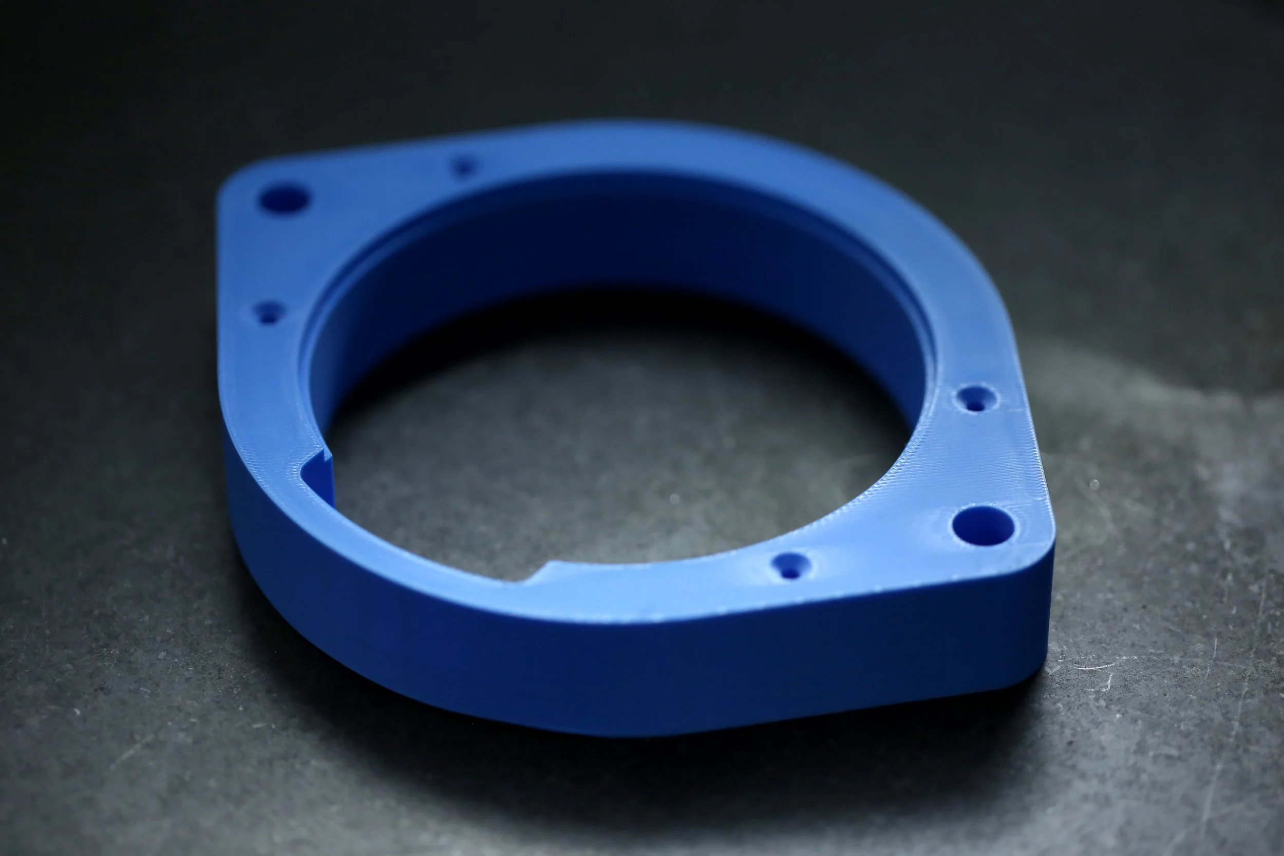 A blue circular plastic component with mounting holes, resting on a dark surface.