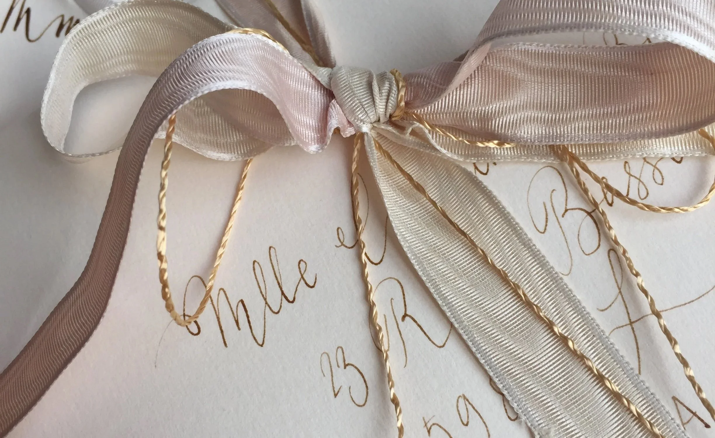 Close-up of elegant envelop with a cream-coloured ribbon tied in a bow, gold accent strings, and gold cursive writing partially visible underneath.