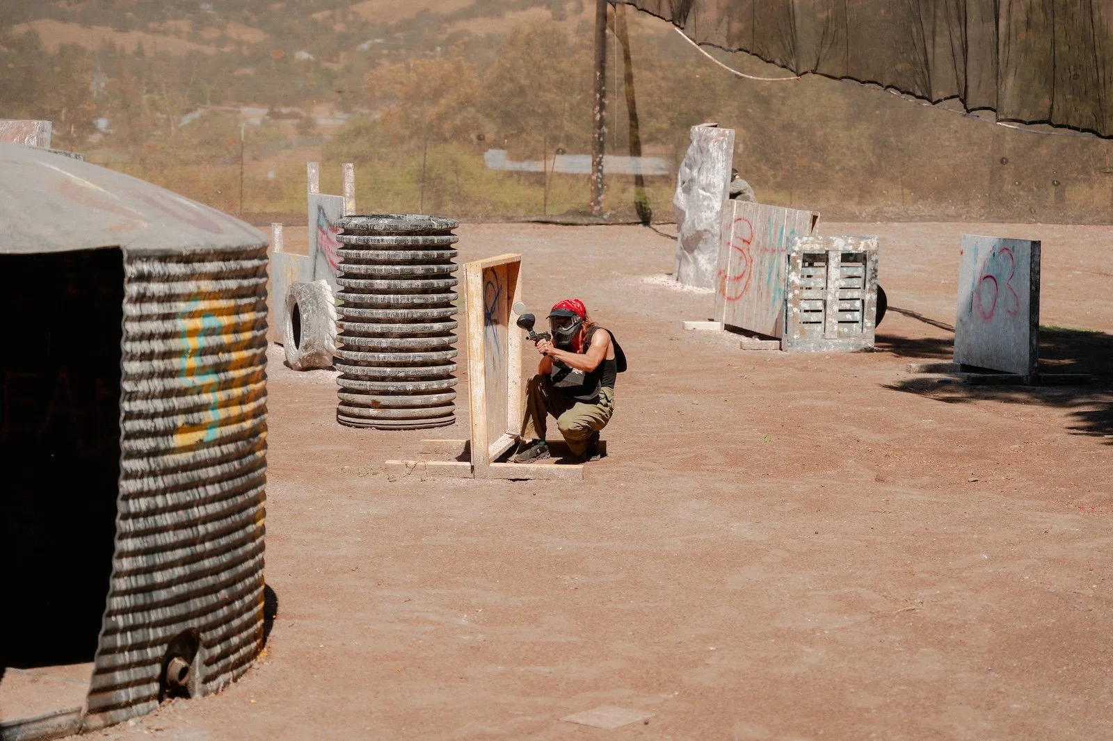 A person in tactical gear participating in a paintball game, crouching behind a wooden barrier on a dirt field with various obstacles and barriers around.