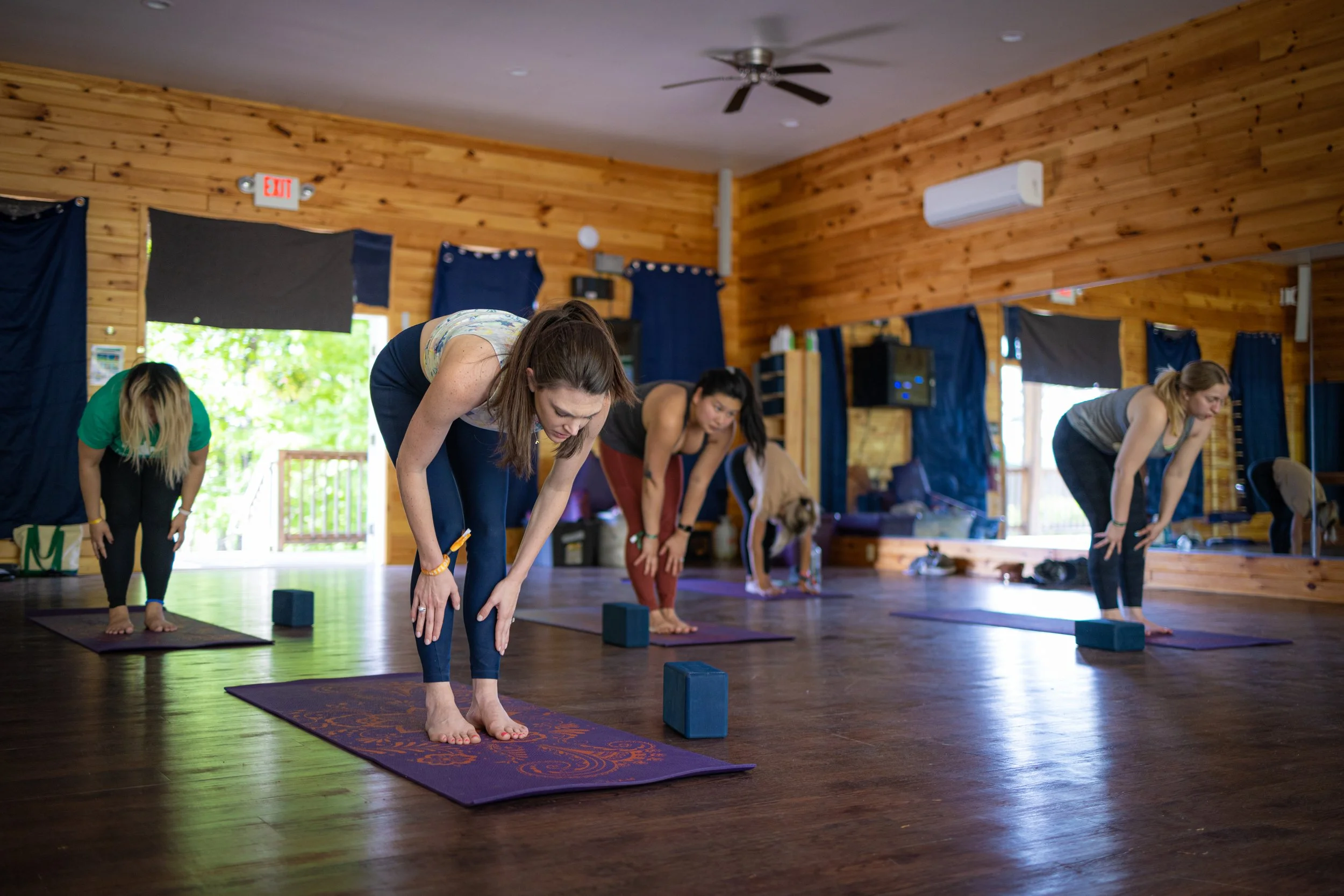 A group of women participating in a yoga class in a wooden-floored studio, practicing standing forward bend pose using yoga blocks for support.