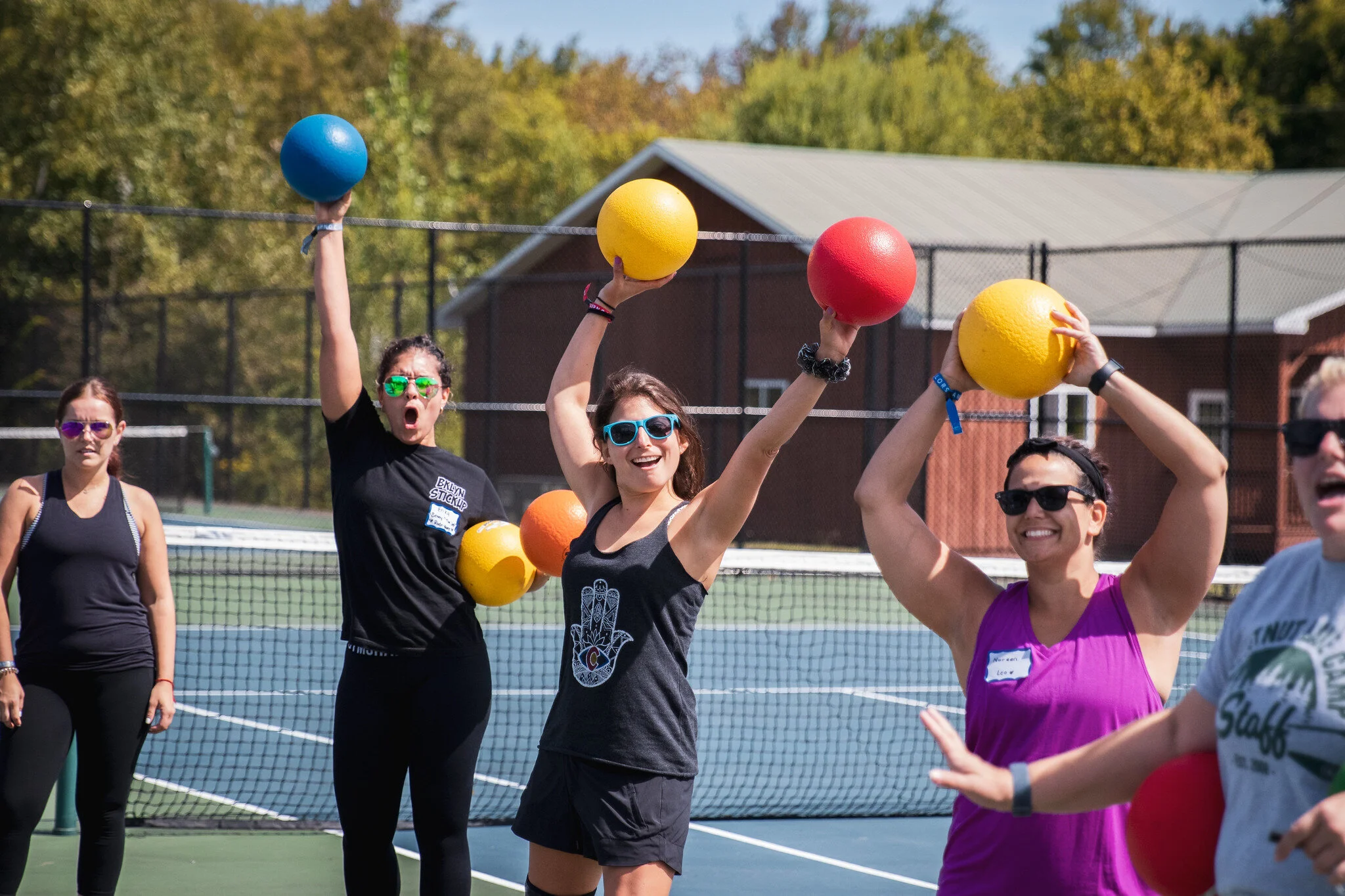 Group of six women playing pickleball outdoors on a sunny day, holding colorful balls, some wearing sunglasses, on a tennis court with trees and a building in the background.