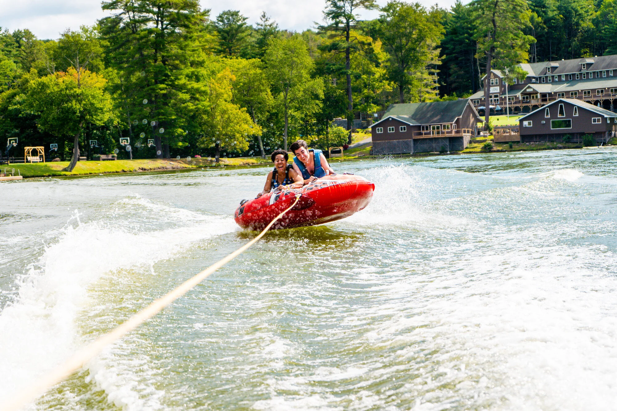 Two people on a red inflatable boat being pulled by a rope on the water, smiling as they hold on. In the background are green trees, houses, and a small dock along the shoreline.