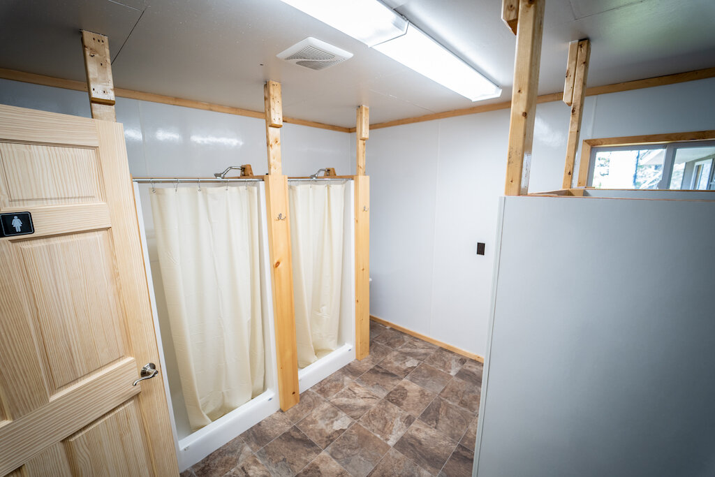 Interior of a bathroom under construction with two shower stalls with curtains, a door with a unisex restroom sign, and a window showing trees outside.