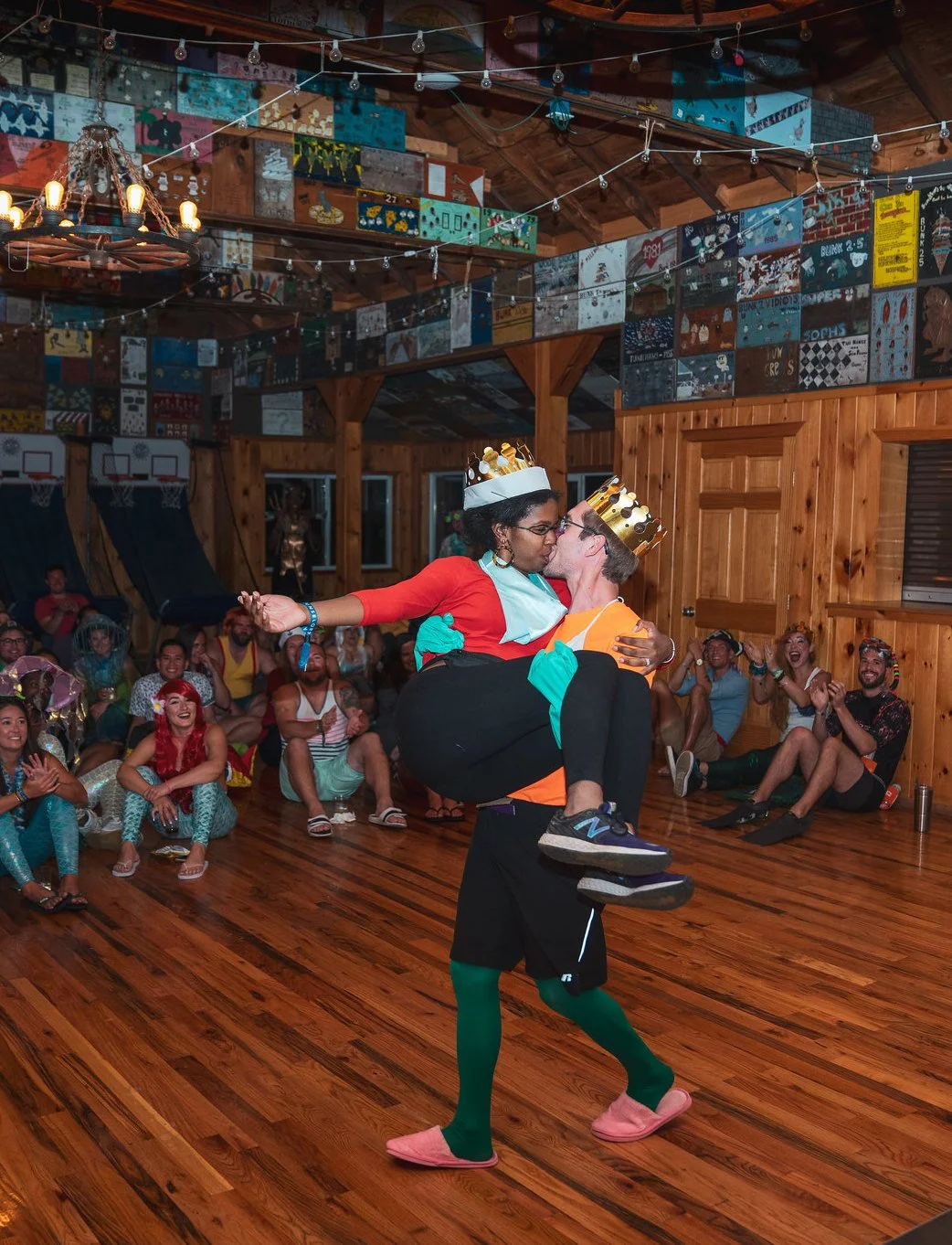 People dressed in costumes celebrating at a party in a wooden hall. A man is carrying a woman while they kiss, both wearing paper crown hats. The onlookers in the background are laughing and clapping, some in colorful costumes.