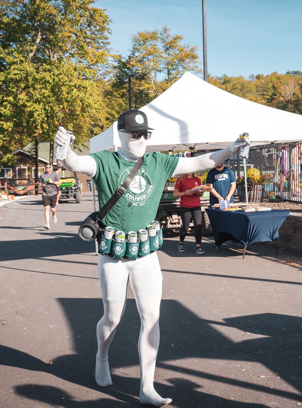Person dressed in a costume resembling a beer can, with a belt of empty cans around their waist, black sunglasses, and wearing a green T-shirt, white tights, and a black baseball cap, standing outdoors at a festival or event with tents and people in the background.