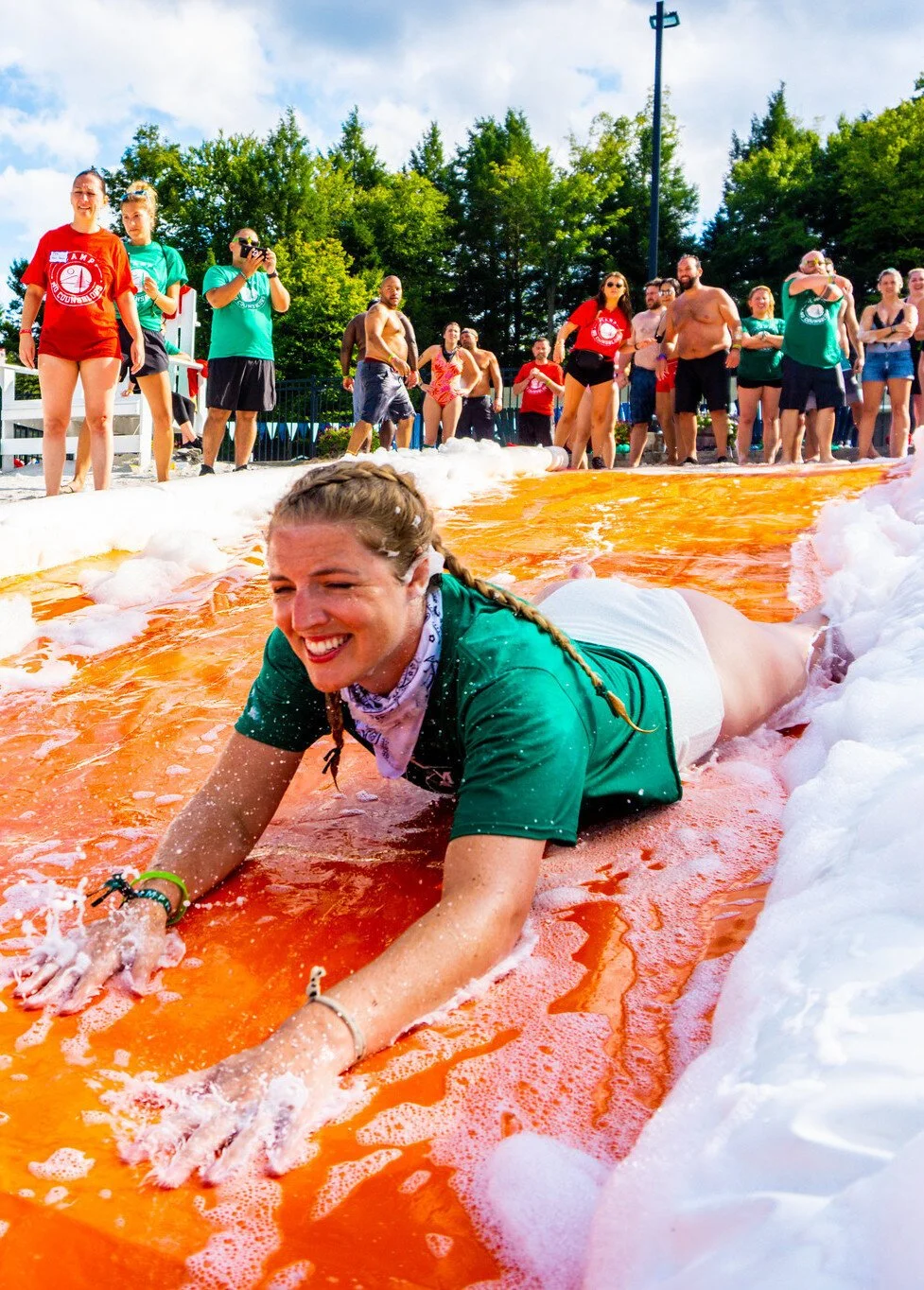 A woman crawling on an orange slip and slide covered in foam, with a big group of people watching behind her at an outdoor event on a sunny day.