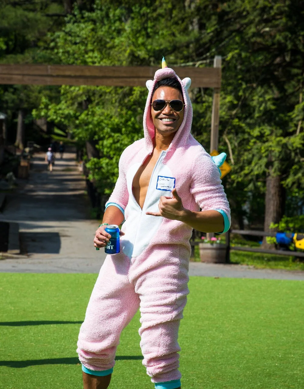 Man in pink unicorn onesie and sunglasses smiling outdoors, holding a soda can and showing a hand sign.