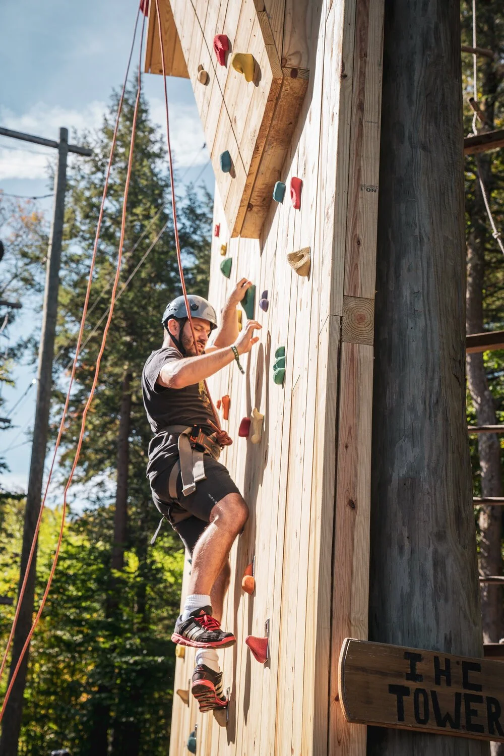 A man wearing a helmet and harness climbing an outdoor wooden rock wall with colorful handholds and footholds, surrounded by trees.