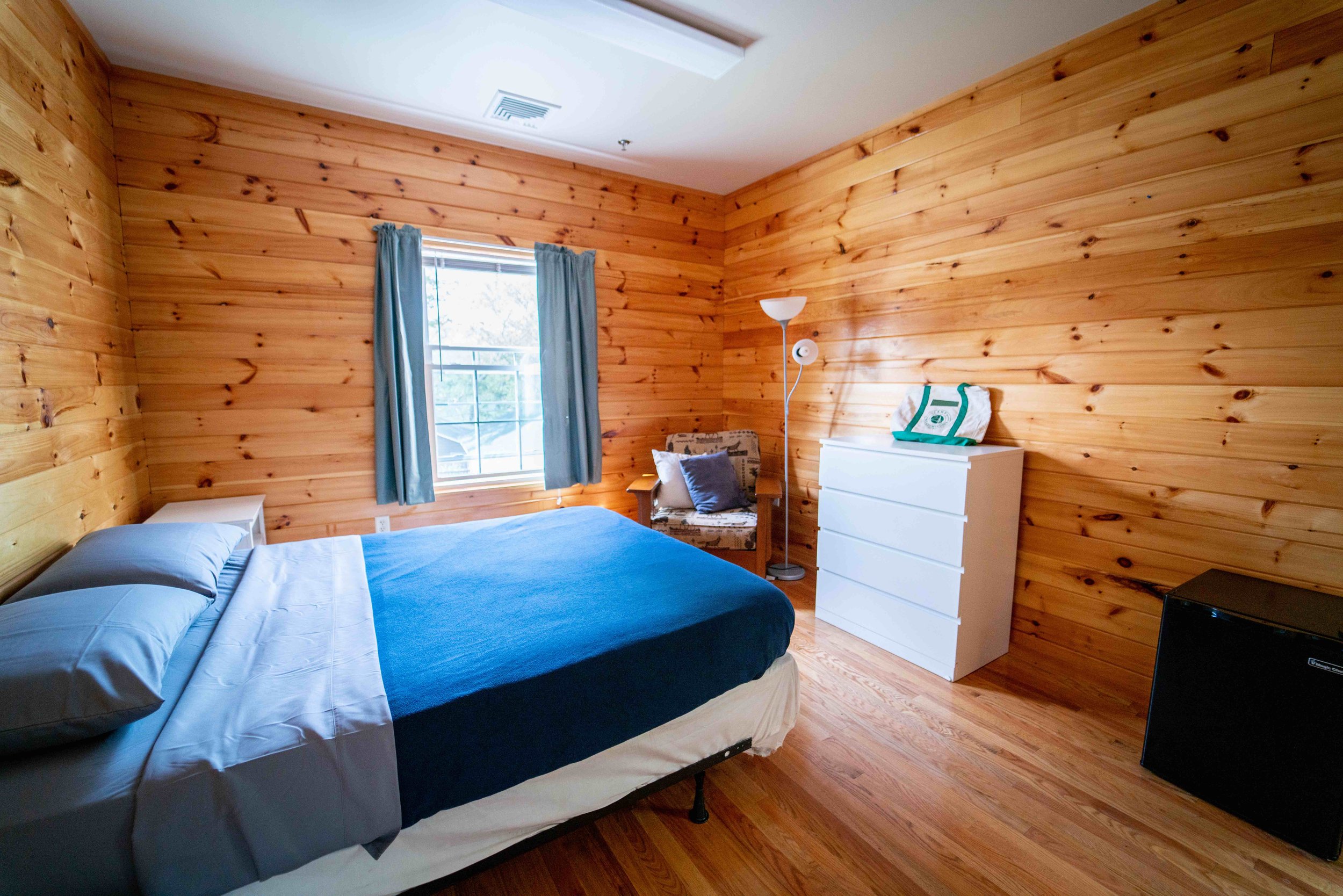 Wood-paneled bedroom with a bed, blue bedspread, gray pillows, a window with light blue curtains, a white dresser, a black mini fridge, a patterned armchair with a dark pillow, a floor lamp, and a small white ceiling vent.