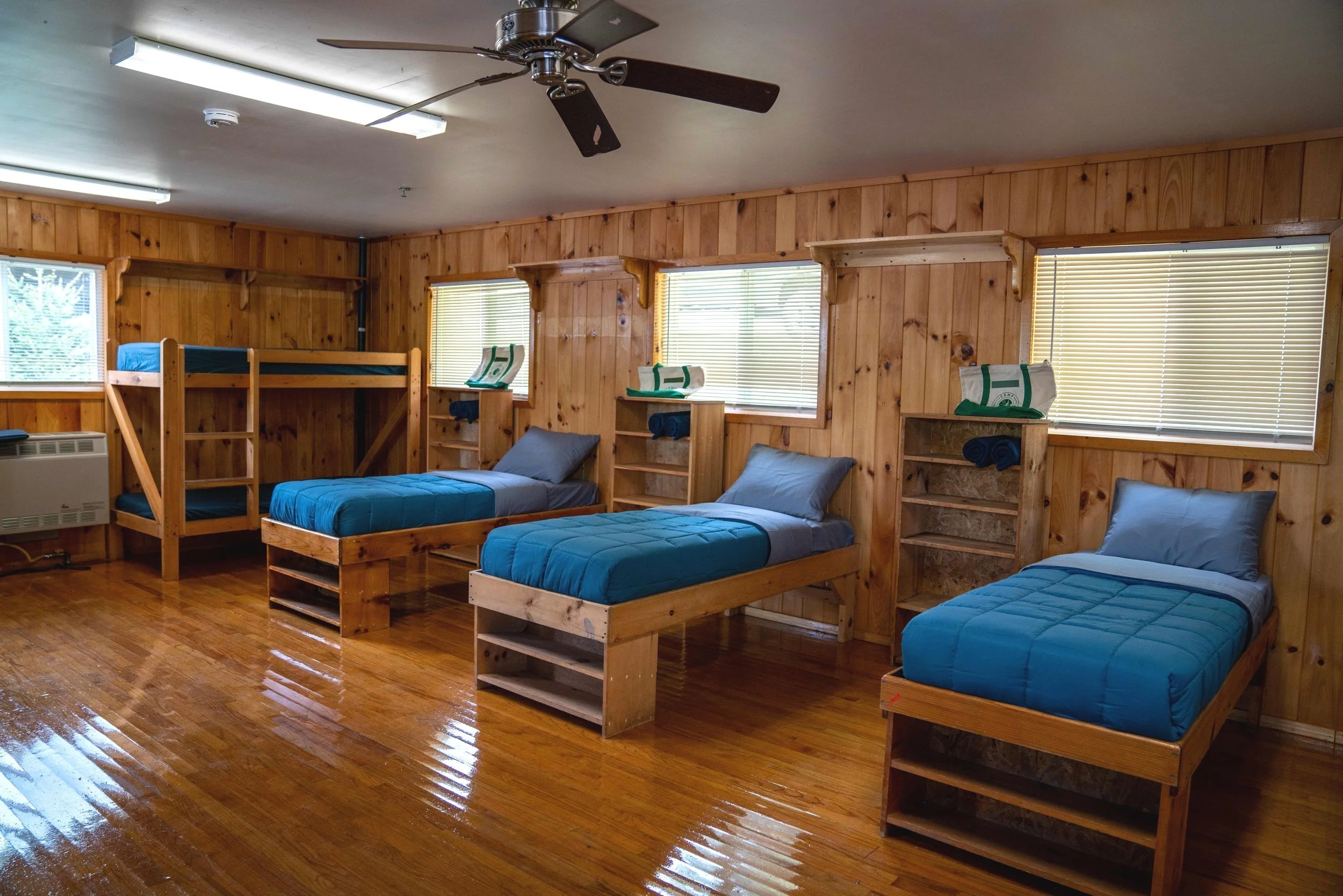 A room with four beds, three single beds with blue bedding and one bunk bed with a wooden frame, all against wooden paneled walls and with blinds-covered windows.