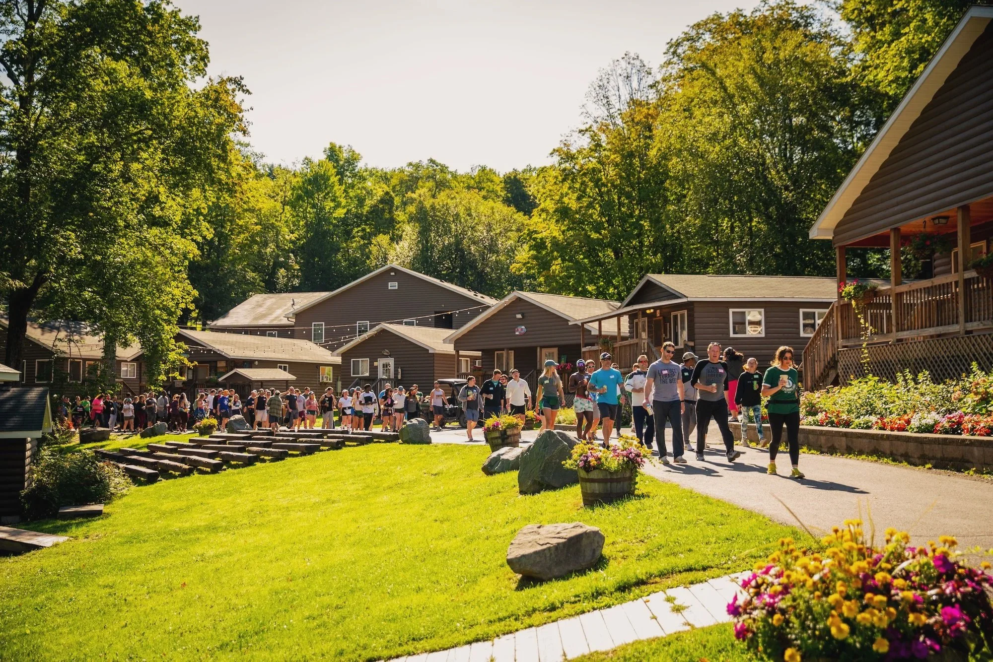 A large group of people walking outdoors on a sunny day in a residential neighborhood with brown houses, green trees, and colorful flowers.