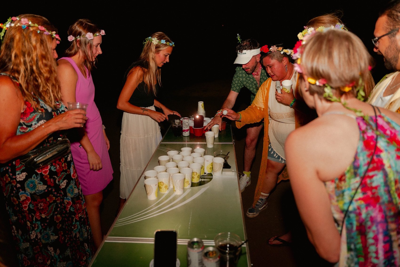 Group of people at a beach party playing a game with ping pong balls and cups around a tabletop, dressed in colorful, tropical attire with floral accessories, during nighttime.